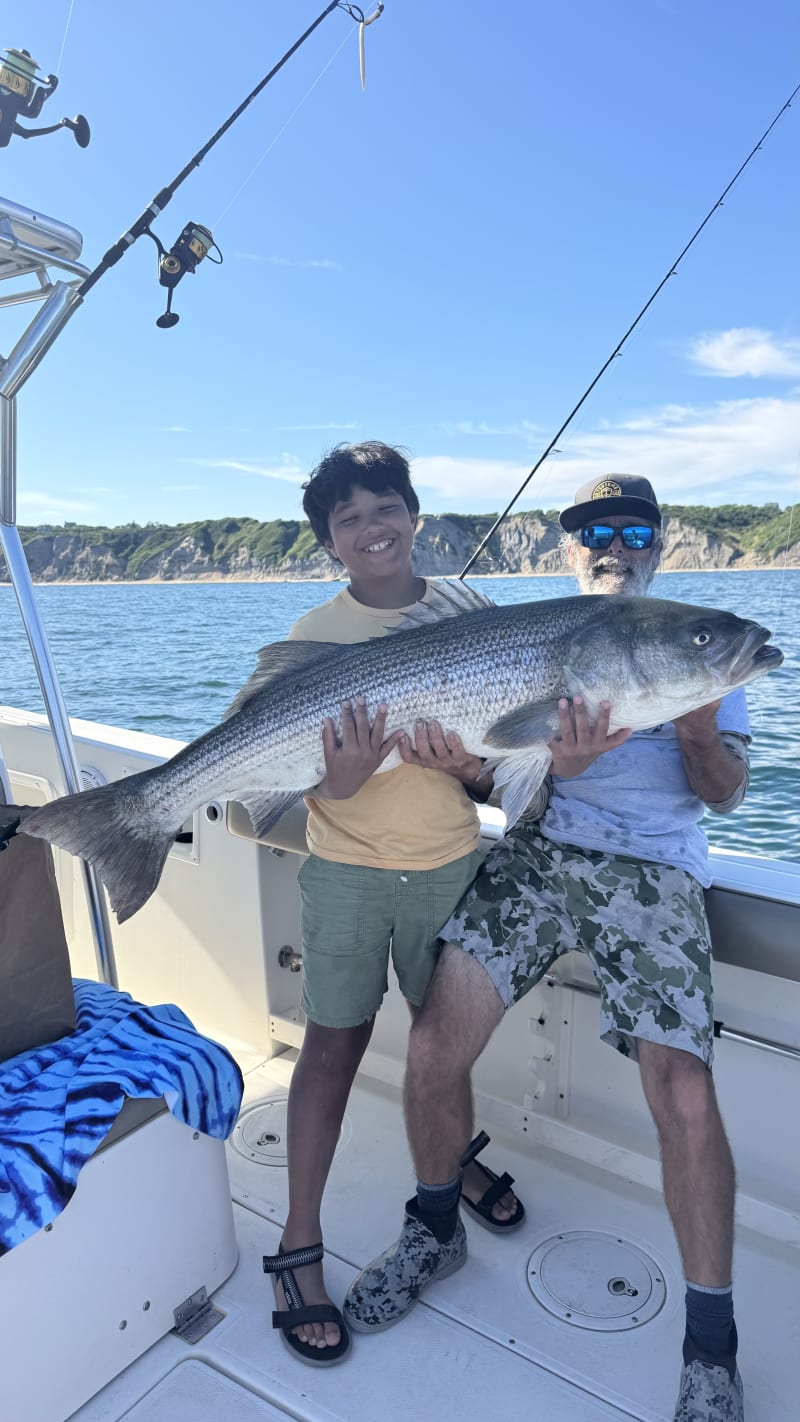 Young angler and Captain Matt holding a striped bass with Block Island cliffs in the background
