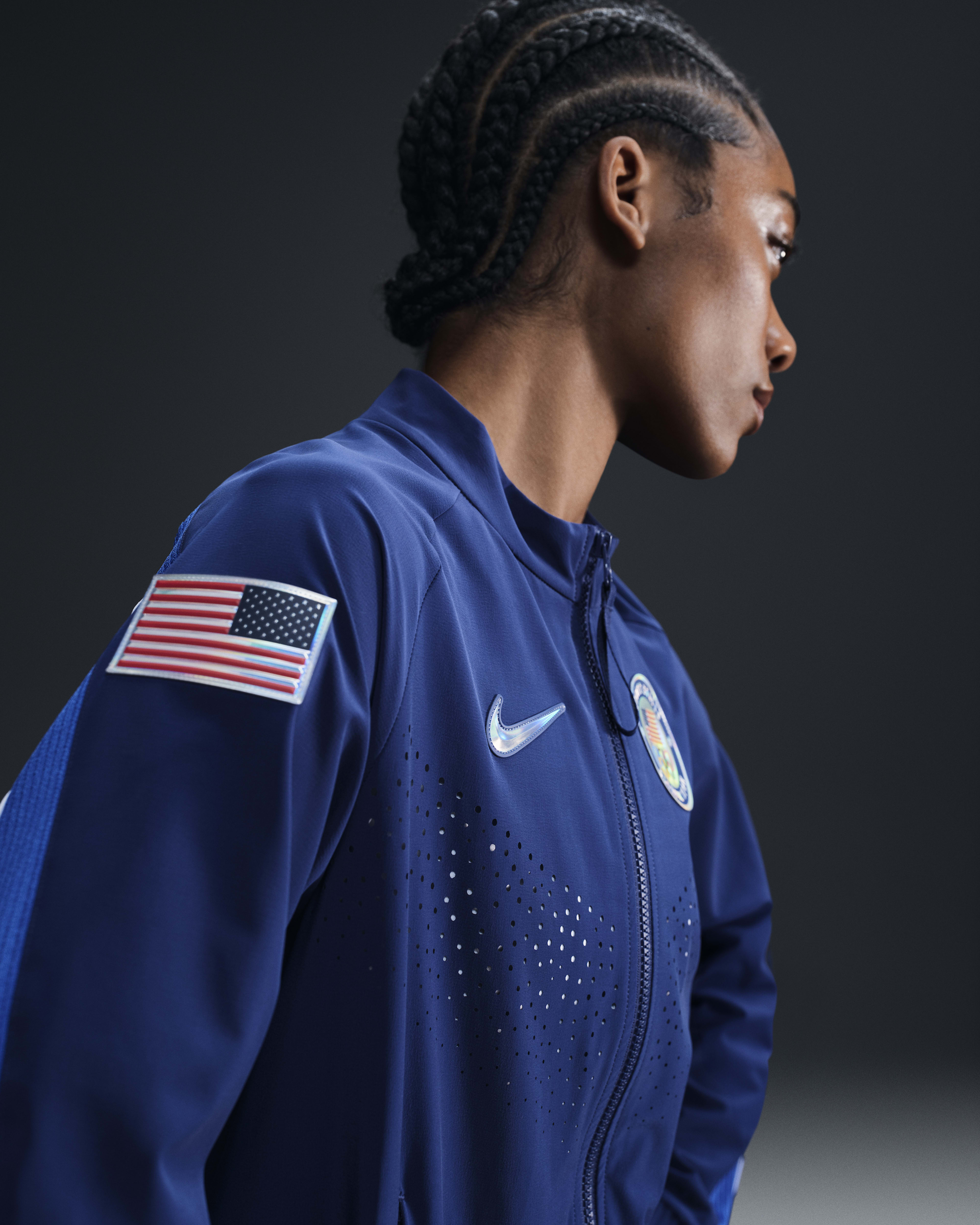 Side view of a female athlete wearing the Women's Medal Ceremony Jacket in the exclusive Team USA bright blue colorway. A USA flag patch is sewn into the upper arm, and the jacket has a silver iridescent Swoosh. The athlete stands against a dark grey background.