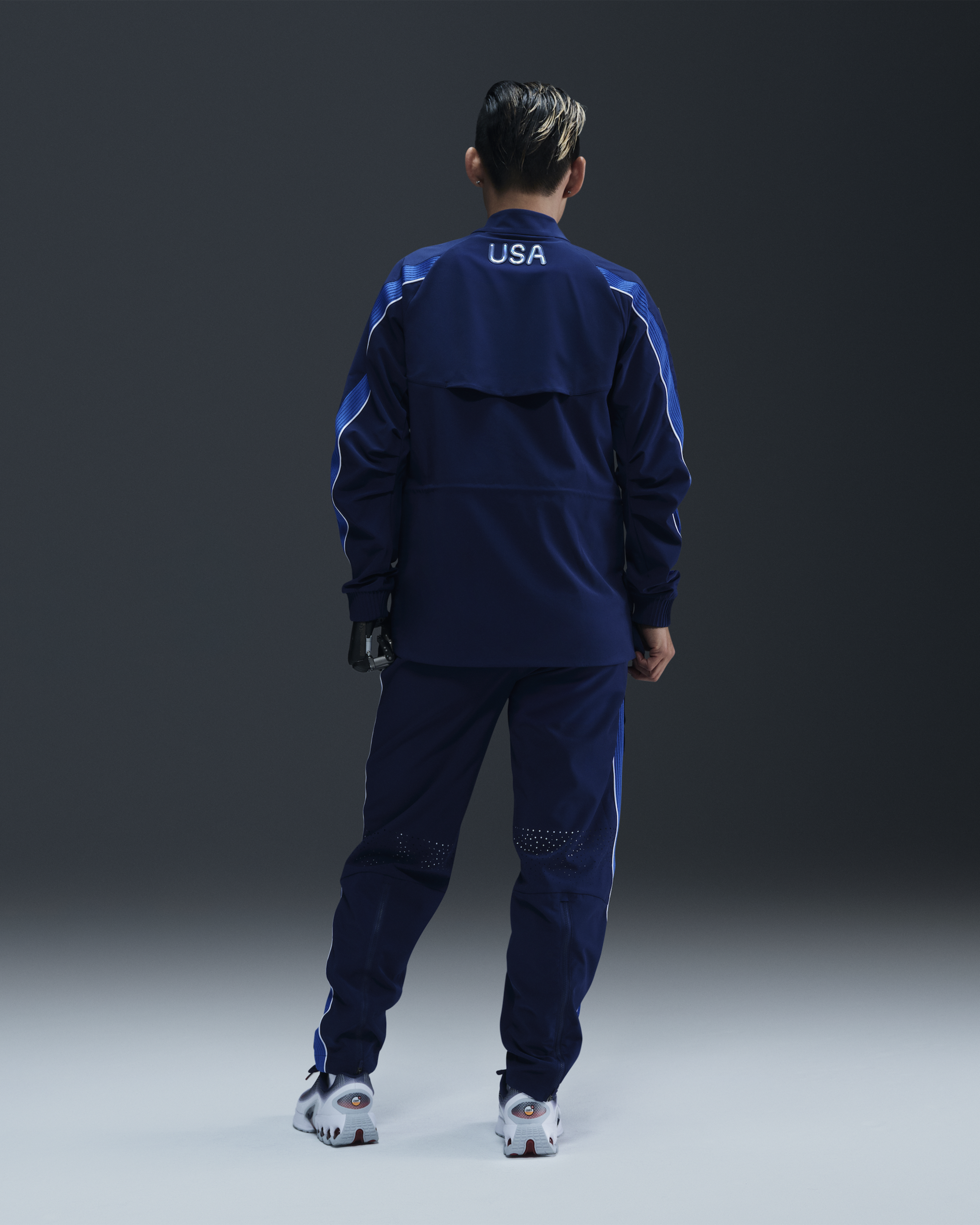 Back view of the full body of a male athlete wearing the Men's Medal Ceremony Jacket and Pant in the exclusive bright blue Team USA colorway. The back of the jacket features USA in iridescent silver letters. The athlete stands on a white floor in the Team USA colorway of the Nike Air Max Dn, against a dark background.