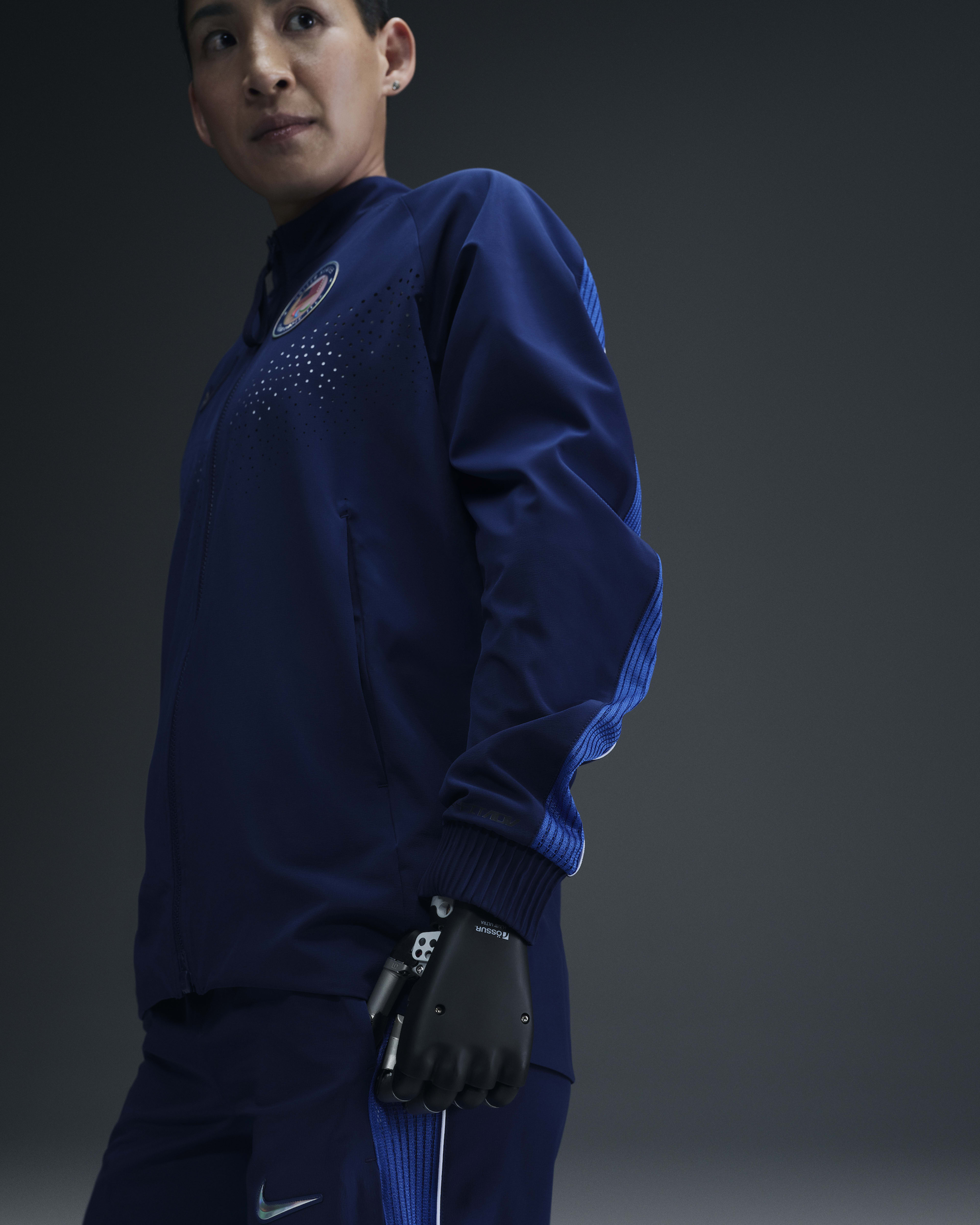 Side view of a female para-athlete, who is wearing the Women's Medal Ceremony Jacket and Pant, in the exclusive bright blue Team USA colorway.  The athlete has a black prosthetic hand and she stands against a grey background.