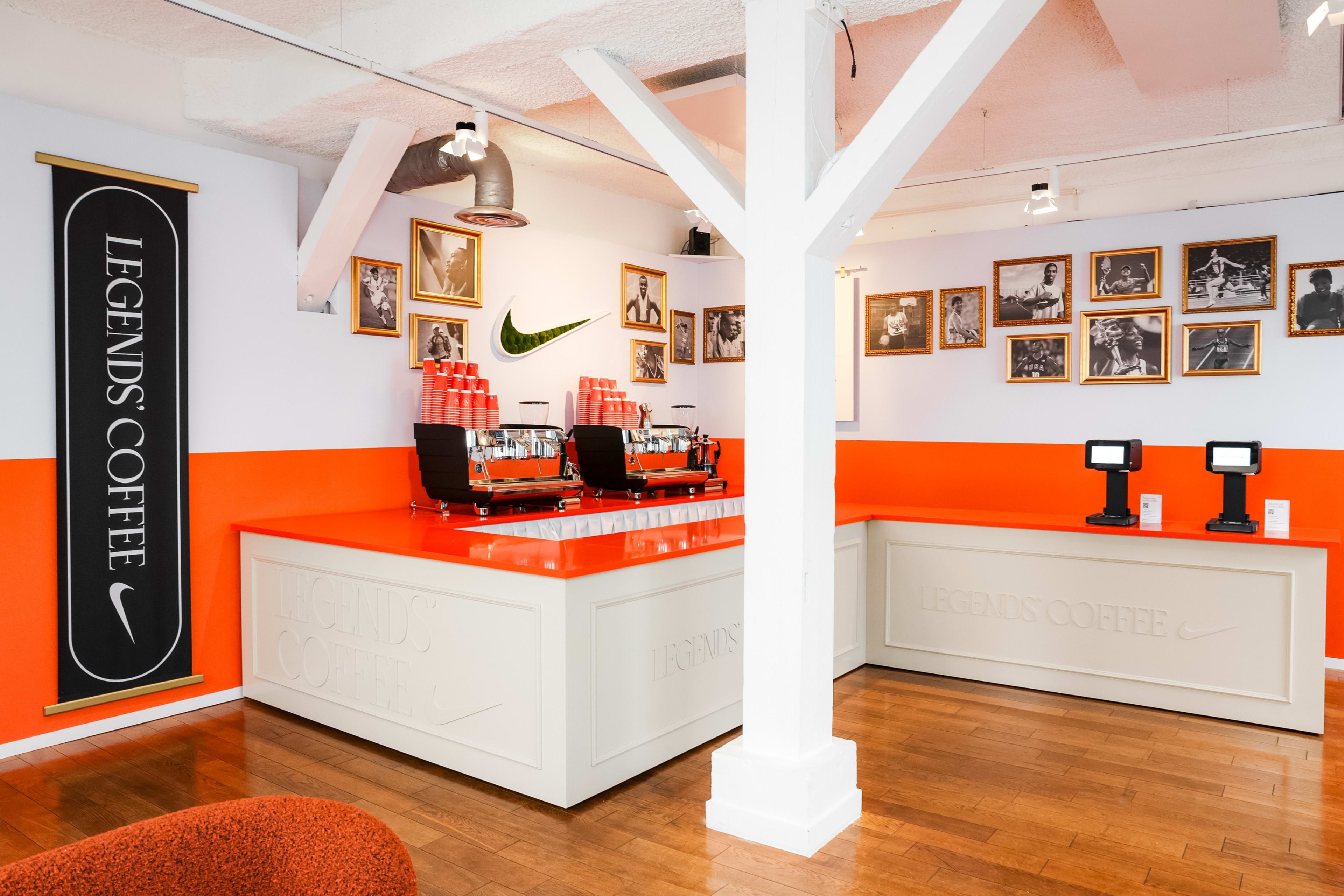 View of the Legends' Coffee bar at the Nike Athlete House in Paris, France. Bright orange and white walls surround the coffee bar, which has a shiny orange counter top. Two silver espresso machines hold stacks of orange coffee cups. Behind the bar are black and white photos of sports lengends.