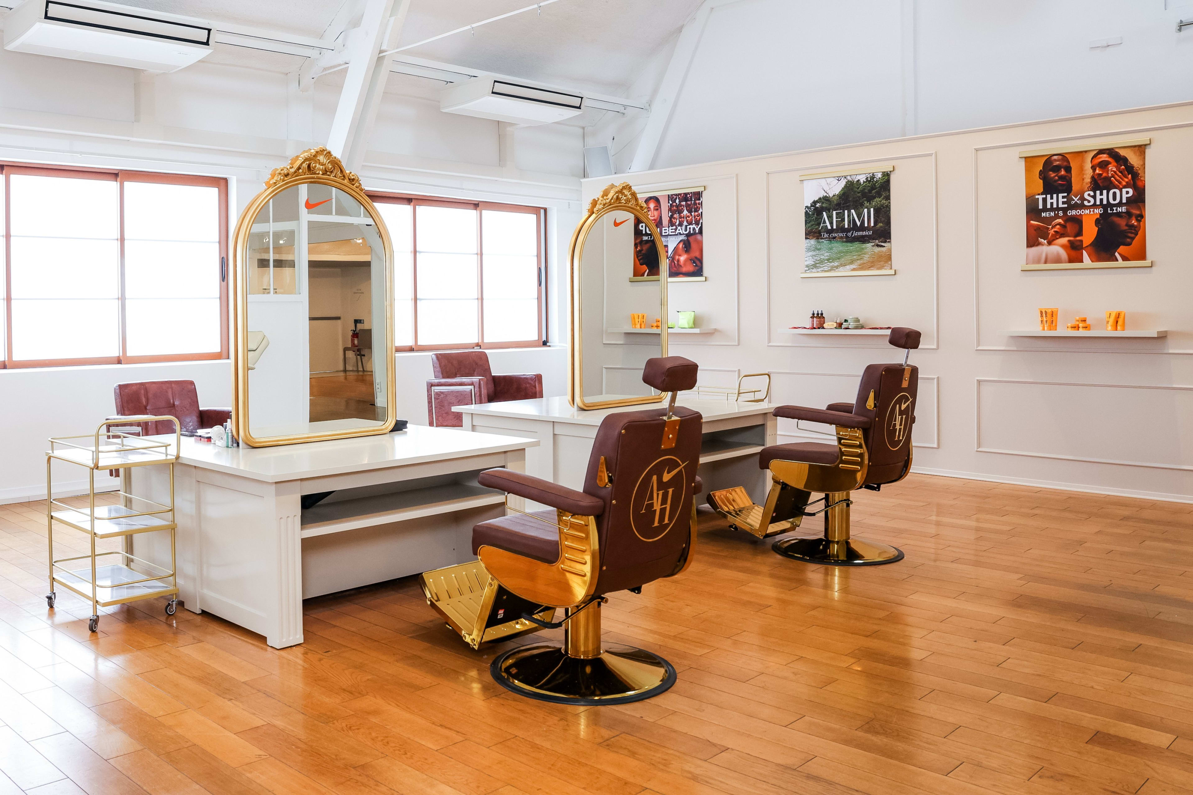 View of the styling bays in the Nike Athlete House in Paris, France. Leather barber chairs with a gold AH logo sit on top of a polished wood floor, in front of gold mirrors and white styling stations. The room is brightly lit with natural light.