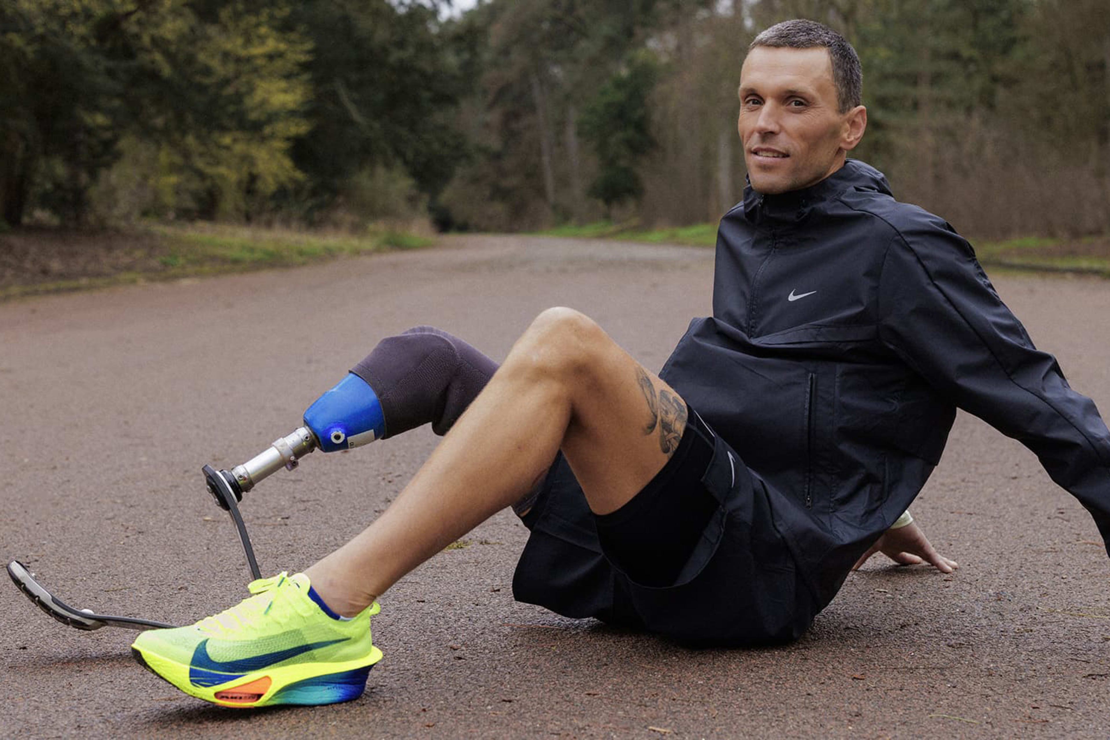 Alex Hanquinquant sits on a forested road smiling towards the camera. Alexis wears black Nike Stride shorts and Nike Miler jacket, plus the Nike Alphafly 3 in bright Volt green, along with his triathlon blade.