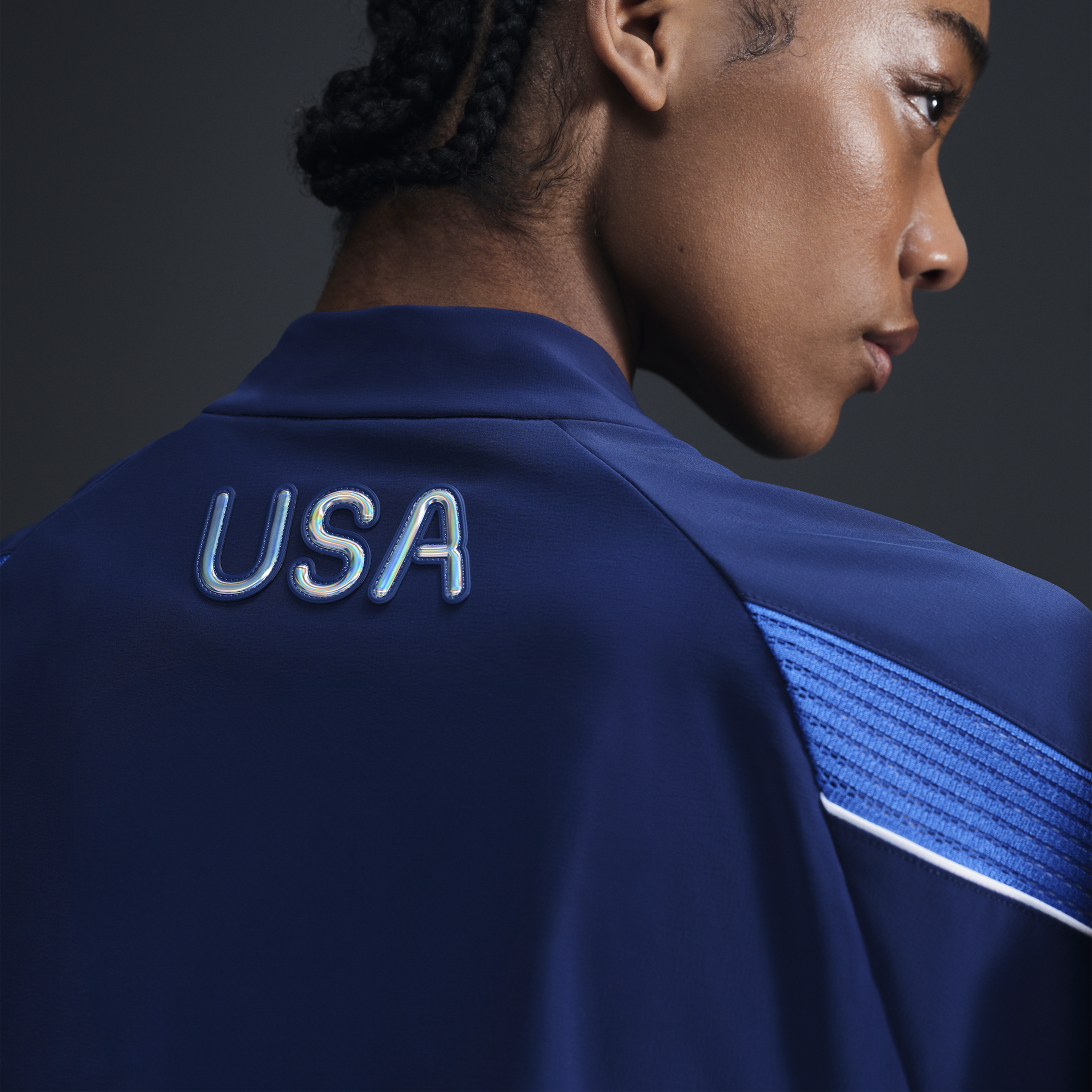 Back view of athlete wearing Women's Medal Ceremony Jacket in the exclusive bright blue Team USA colorway.  USA is emblazoned on the back of the jacket in iridescent silver letters. She stands against a dark background.