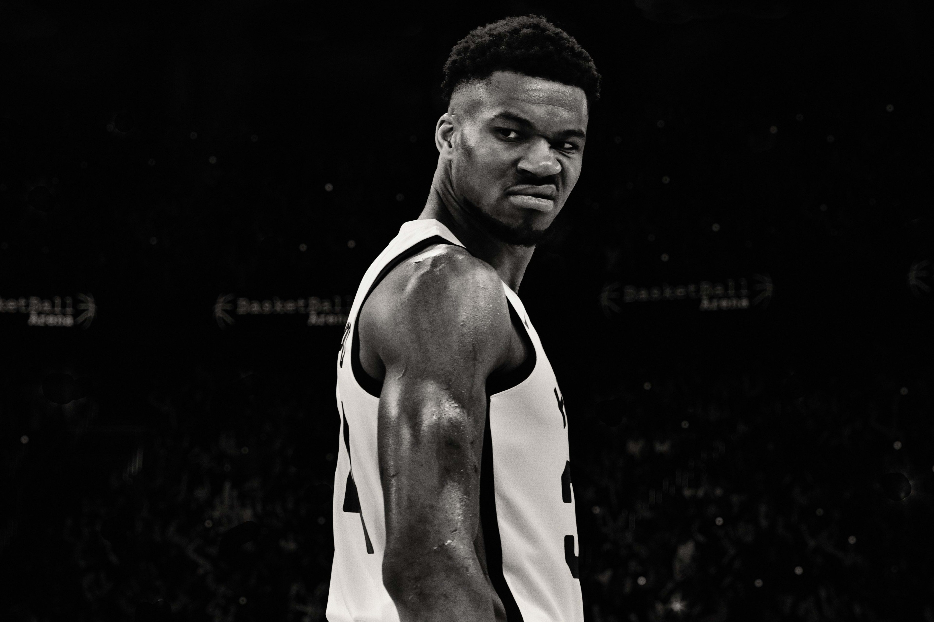 Black and white side angled view of Giannis Antetokounmpo looking over his shoulder at a basketball stadium.