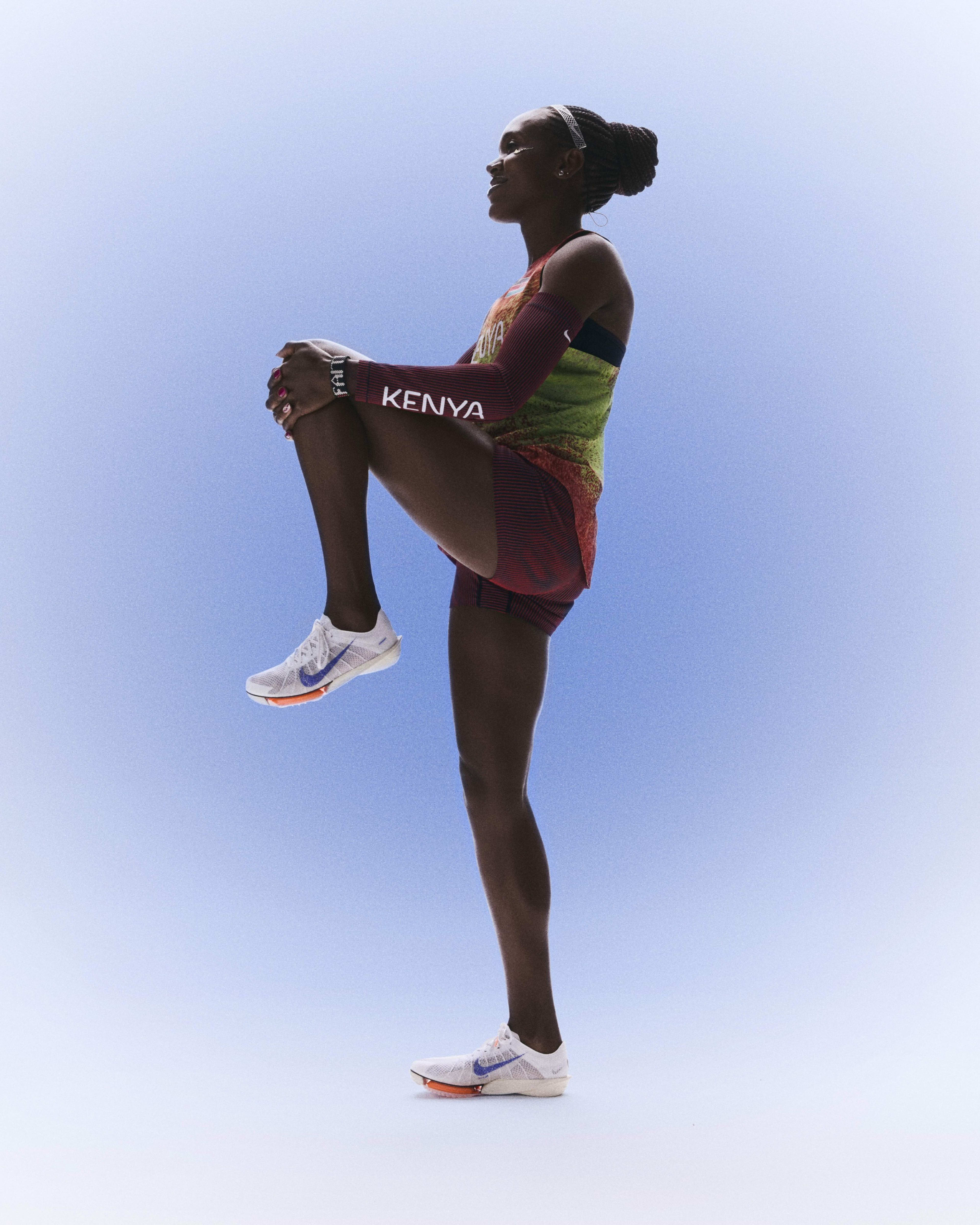 Left view of Faith Kipyegon stretching her left leg by bending it upwards while wearing her Kenya track and field kit uniform.