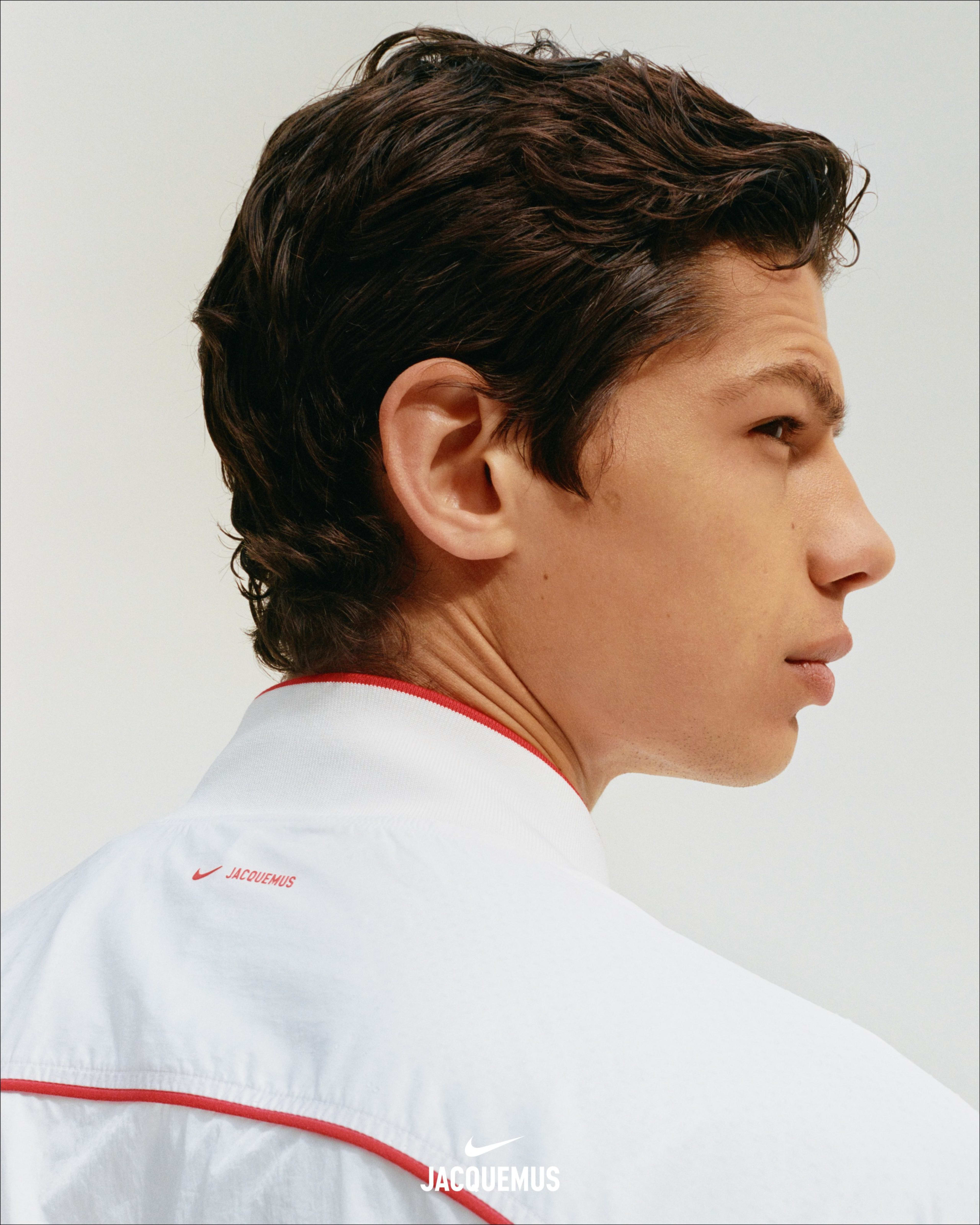 Close up head shot of a male athlete wearing the Nike x Jacquemus Track Jacket in white with university red piping at neckline and across back. We see a red Nike Swoosh and Jacquemas logos.  The man stands against a white background and looks to the right.