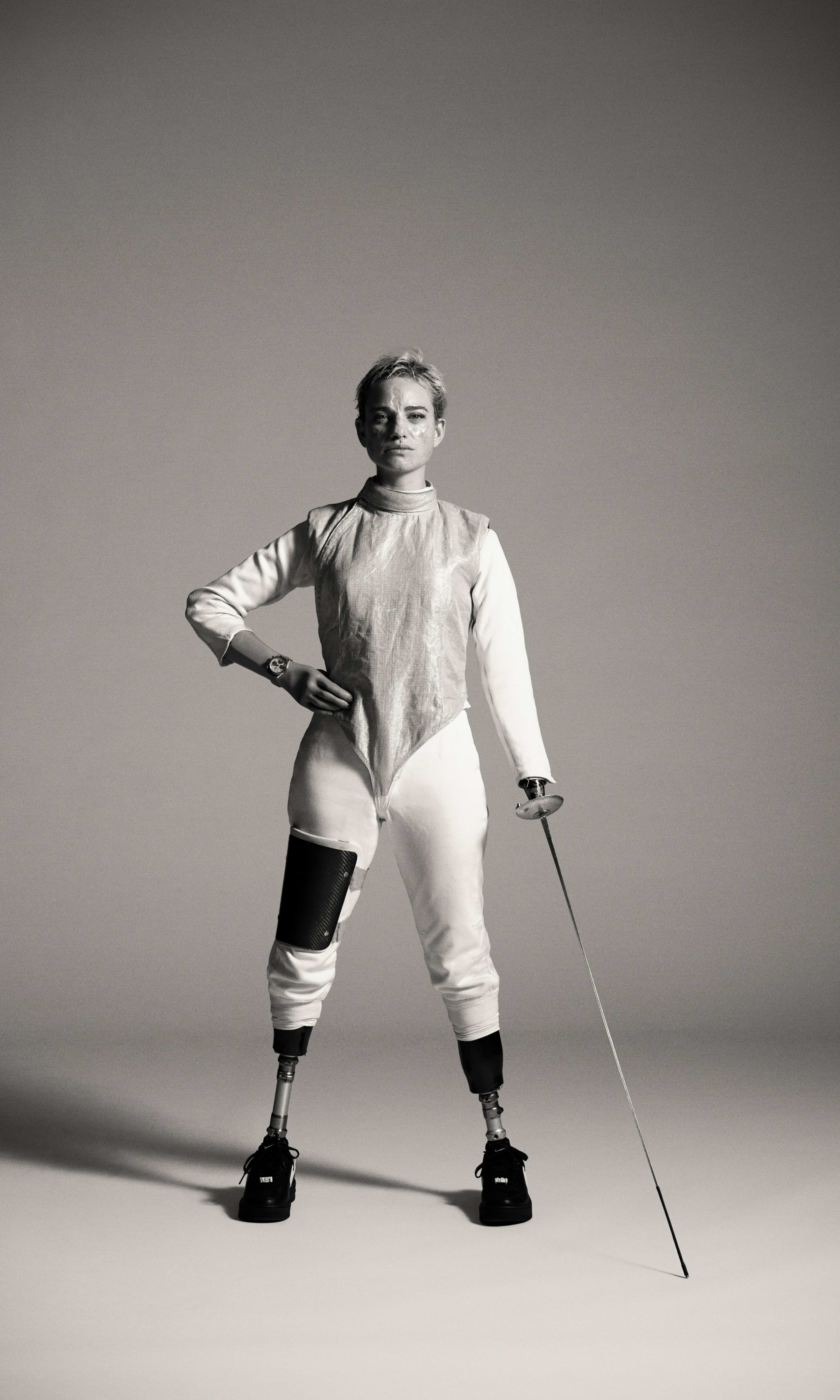 Black and white full body portrait of Bebe Vio, holding one hand to her hip and using her other arm to touch her fencing foil tip to the ground. Bebe looks unsmiling directly at the camera, as she stands in her fencing suit, standing on two prosthetic legs.