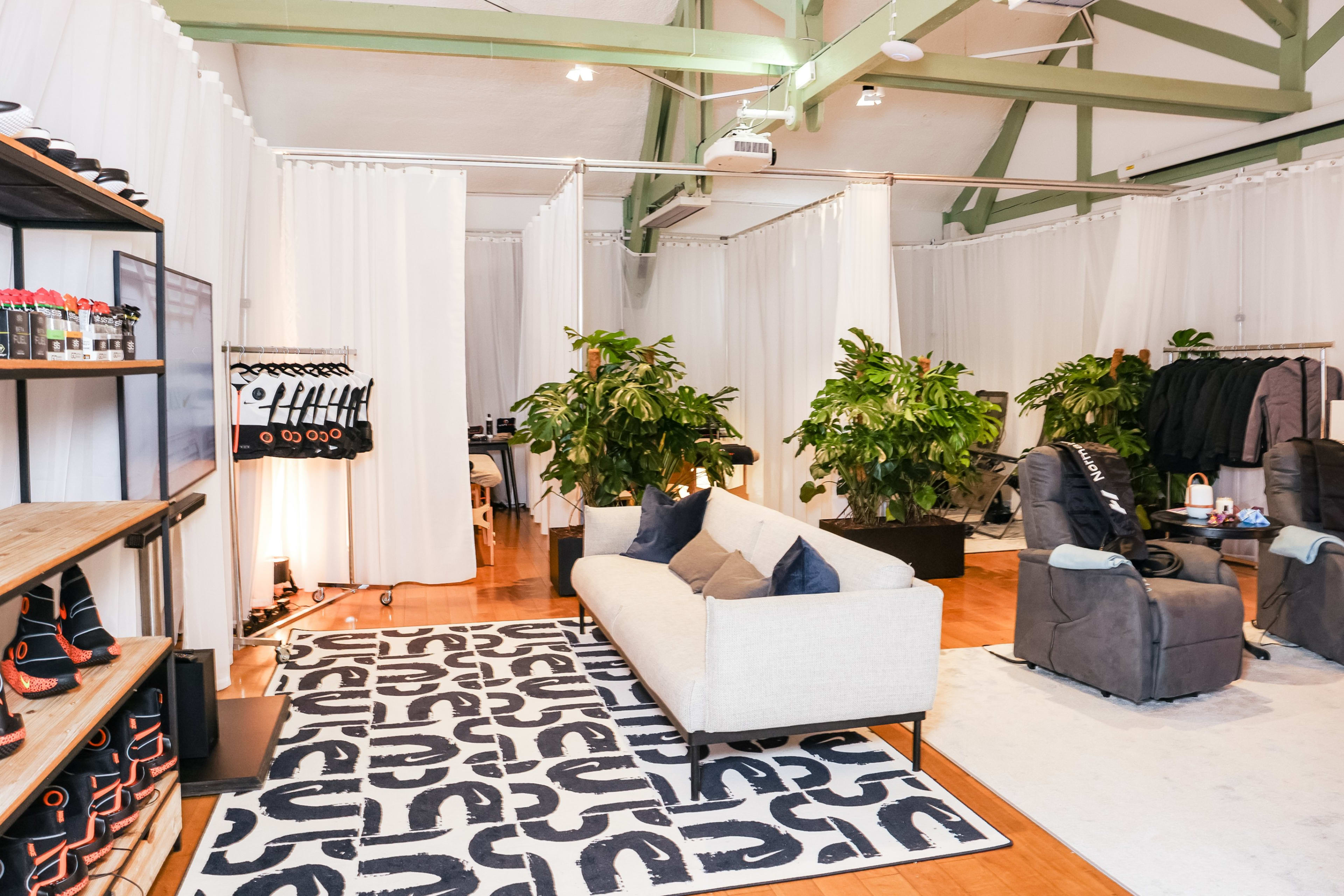 View into the recovery space at the Nike Athlete House in Paris, France. We see a white sofa positioned on a boldly patterned black and white rug. There is a selection of Hyperice vests and boots, as well as massage spaces. White curtained walls surround the area.