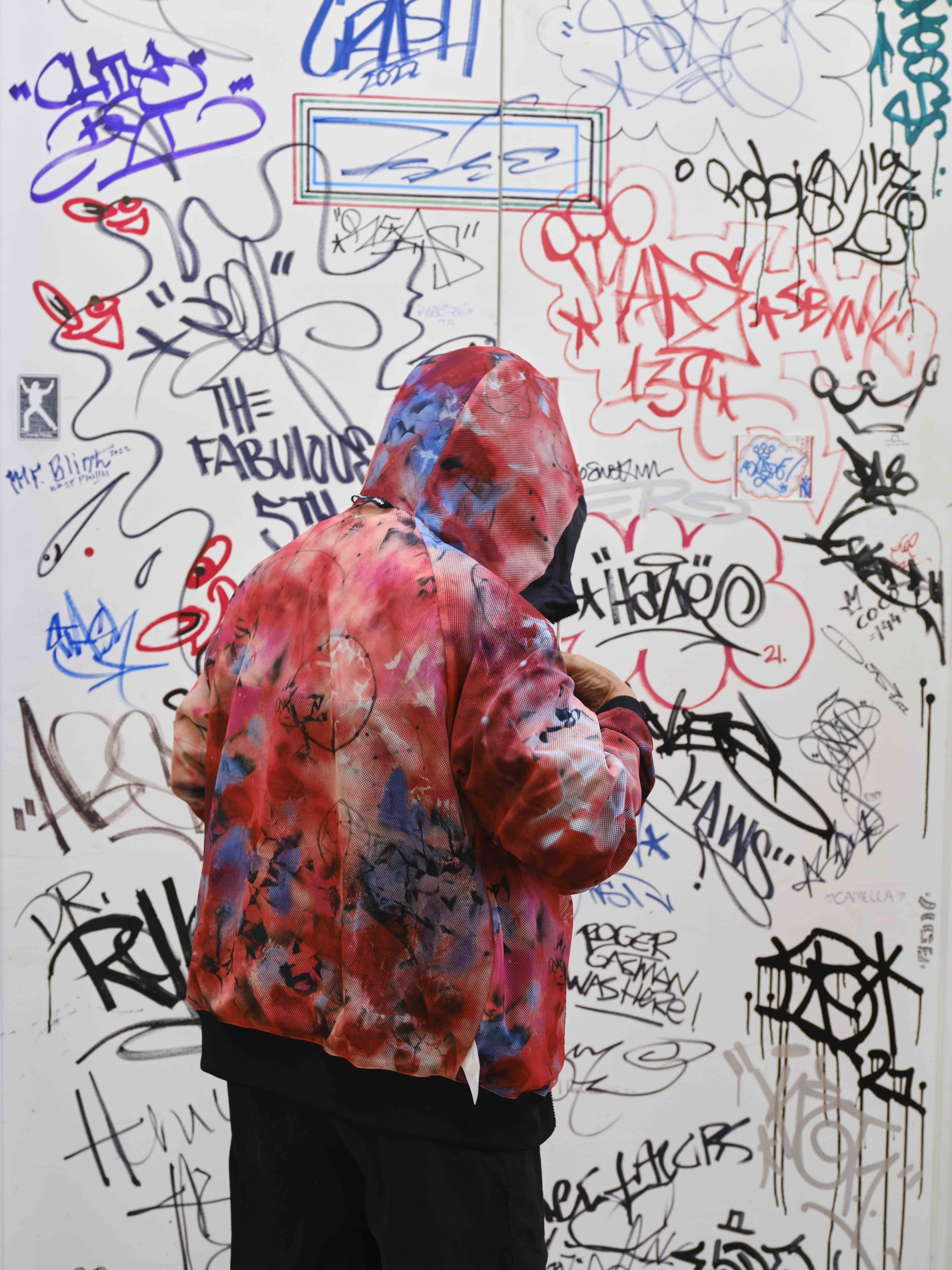 Futura wears a Nike X Futura hoodie featured in Nike's new breaking collection. He wears the piece inside out, showcasing the bright pink, red, and purple liner inspired by artwork, his studio's floor splatter, and his atoms and characters signature. Future stands in front of a white wall covered in colorful graffiti tags.