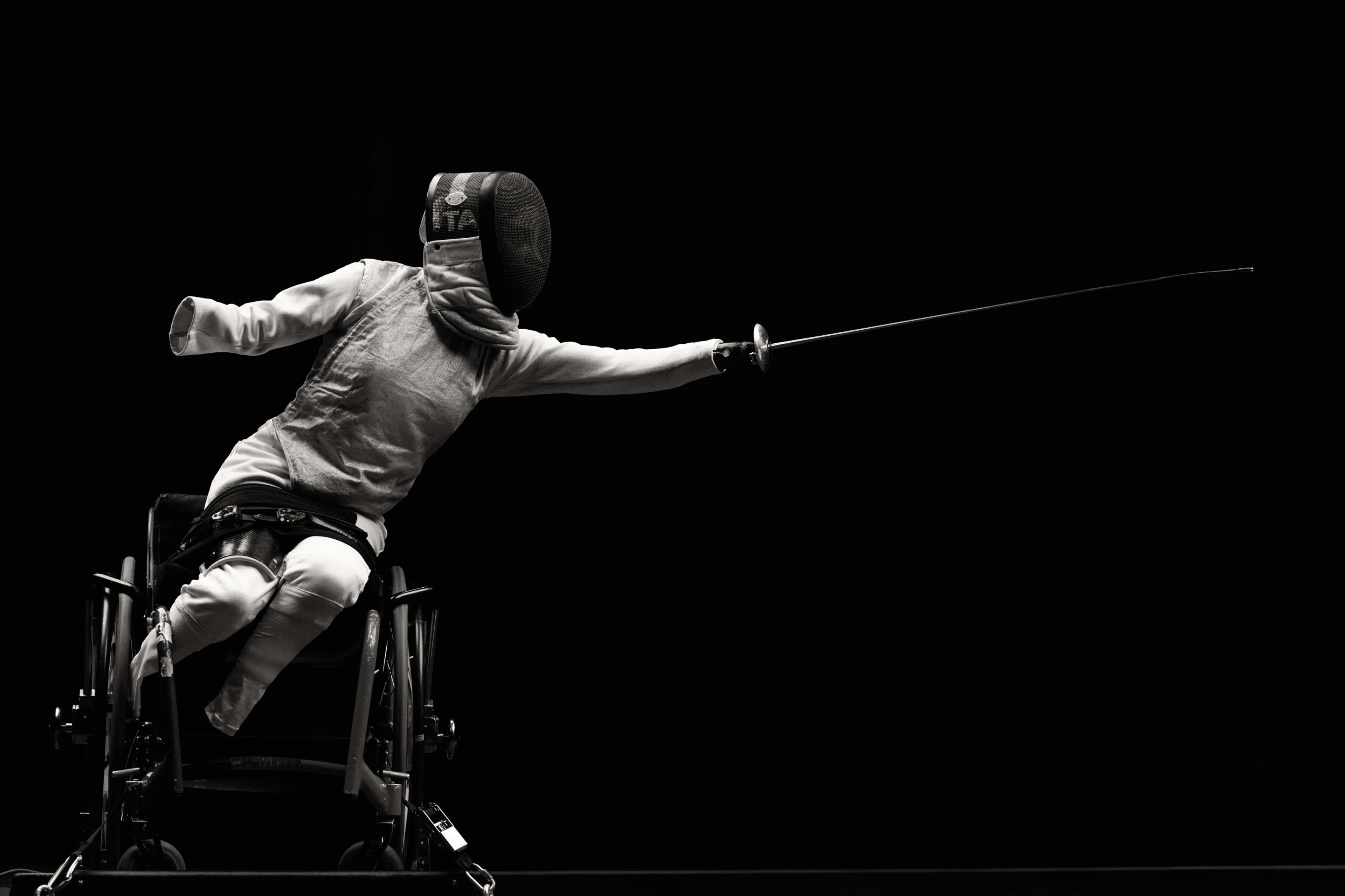 Black and white portrait of Bebe Vio in full fencing uniform and helmet, thrusting her foil to the right. Bebe sits in her performance wheelchair leaning into the thrust with dramatic light shining down on her, against a black background.