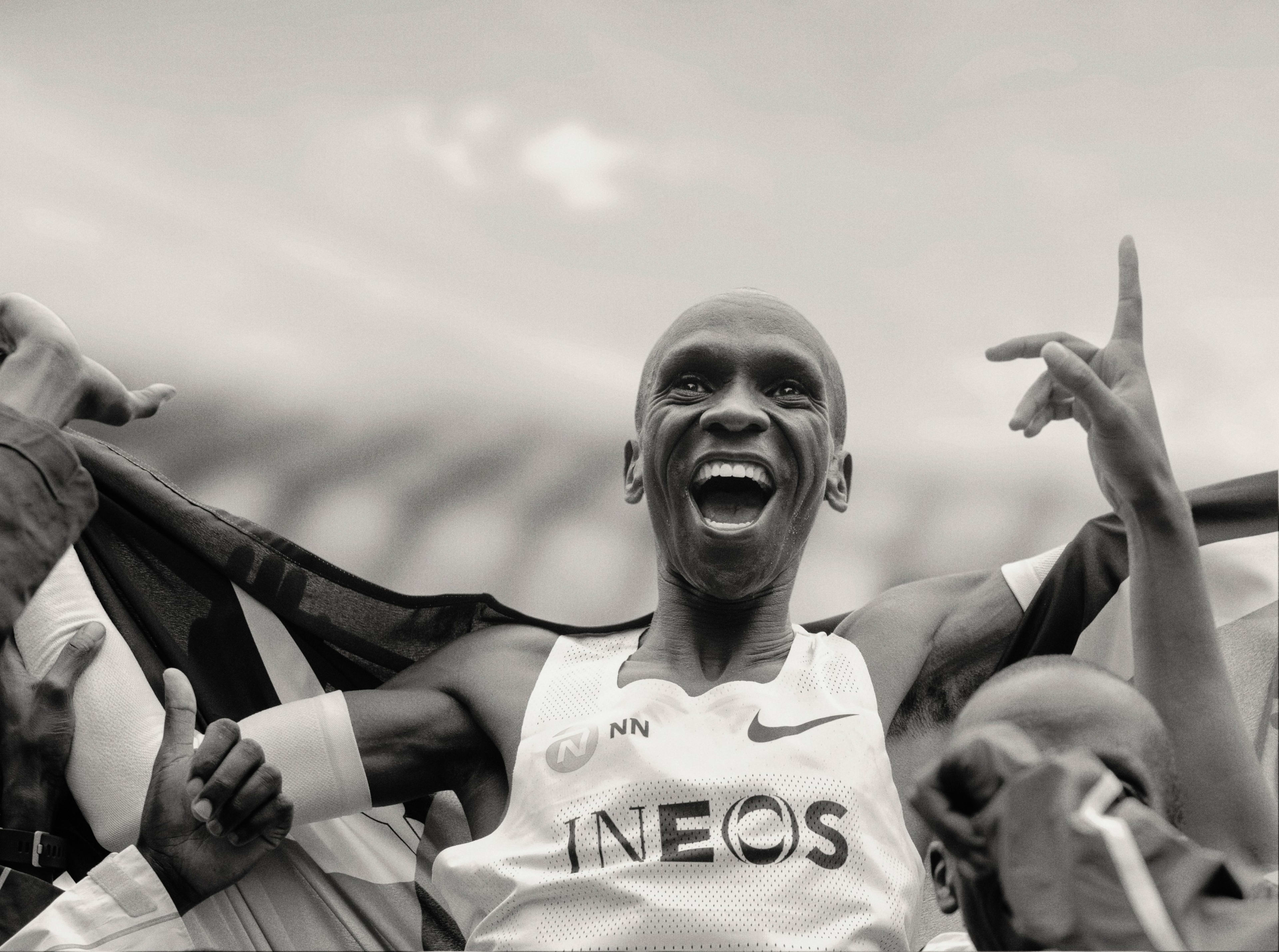 Black and white photo of Eliud Kipchoge smiling widely as he is lifted by a crowd of people.