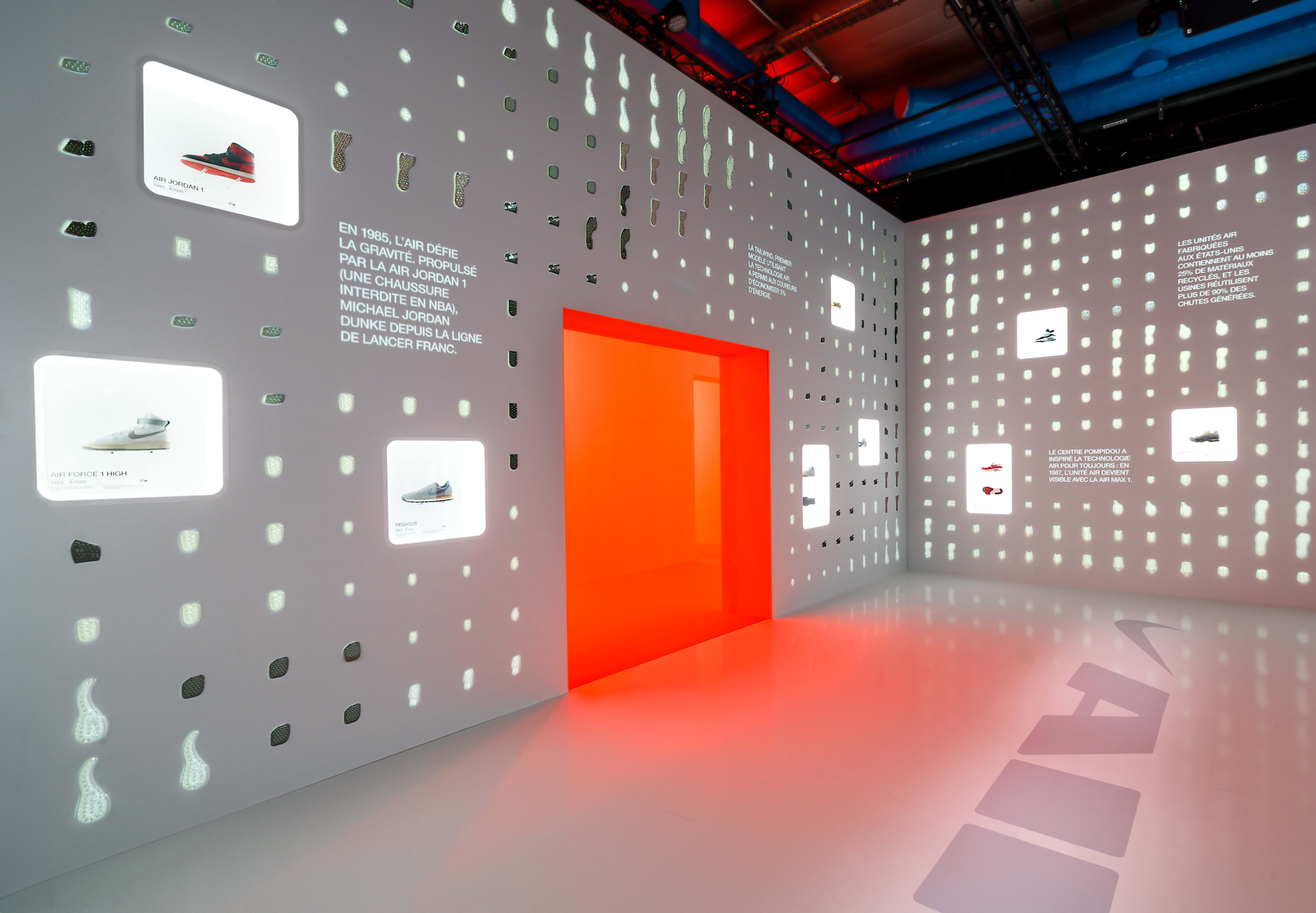 Inside of Gallery 4, Art of Victory exhibity, within the Nike Au Centre Pompidou in Paris, France. Grey walls, backlit with white lighting, display Air units and Nike footwear featuring Air technology. A glowing orange door leads to another exhibit room.