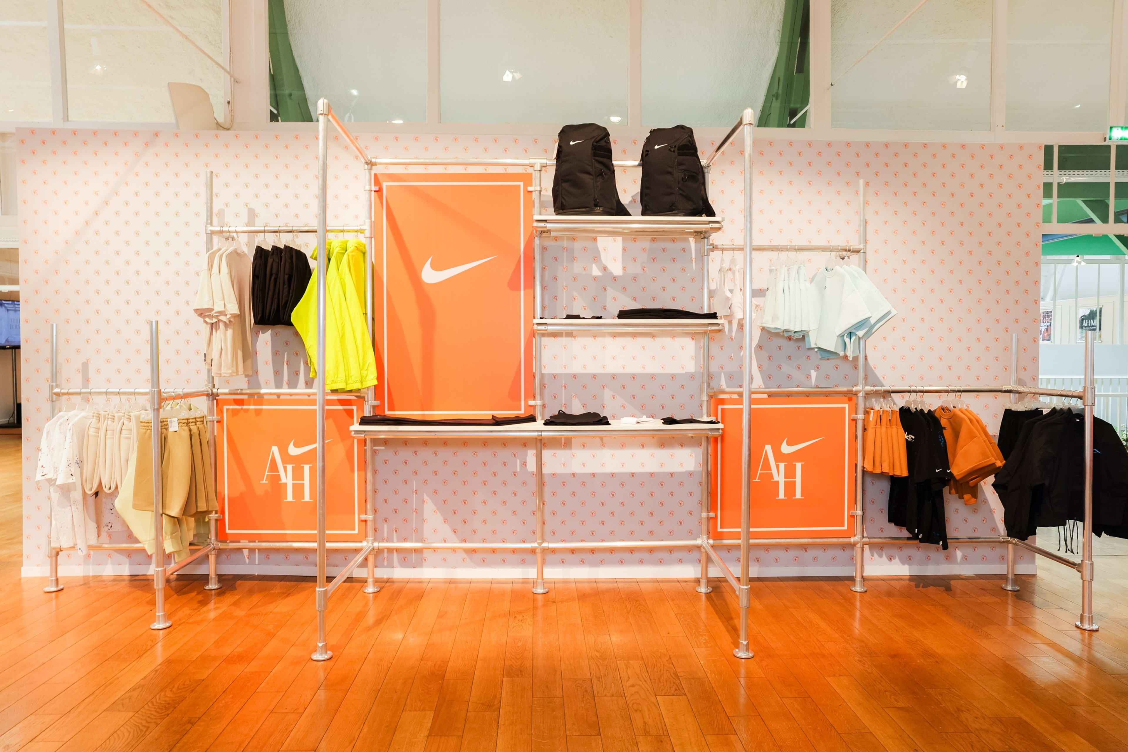 View of the shopping area in the Nike Athletes House in Paris, France. A spectrum of Nike apparel and backpacks are displayed on a silver and white shelving system. The display sits on a polished wood floor and against a wall with a repeating AH logo pattern.