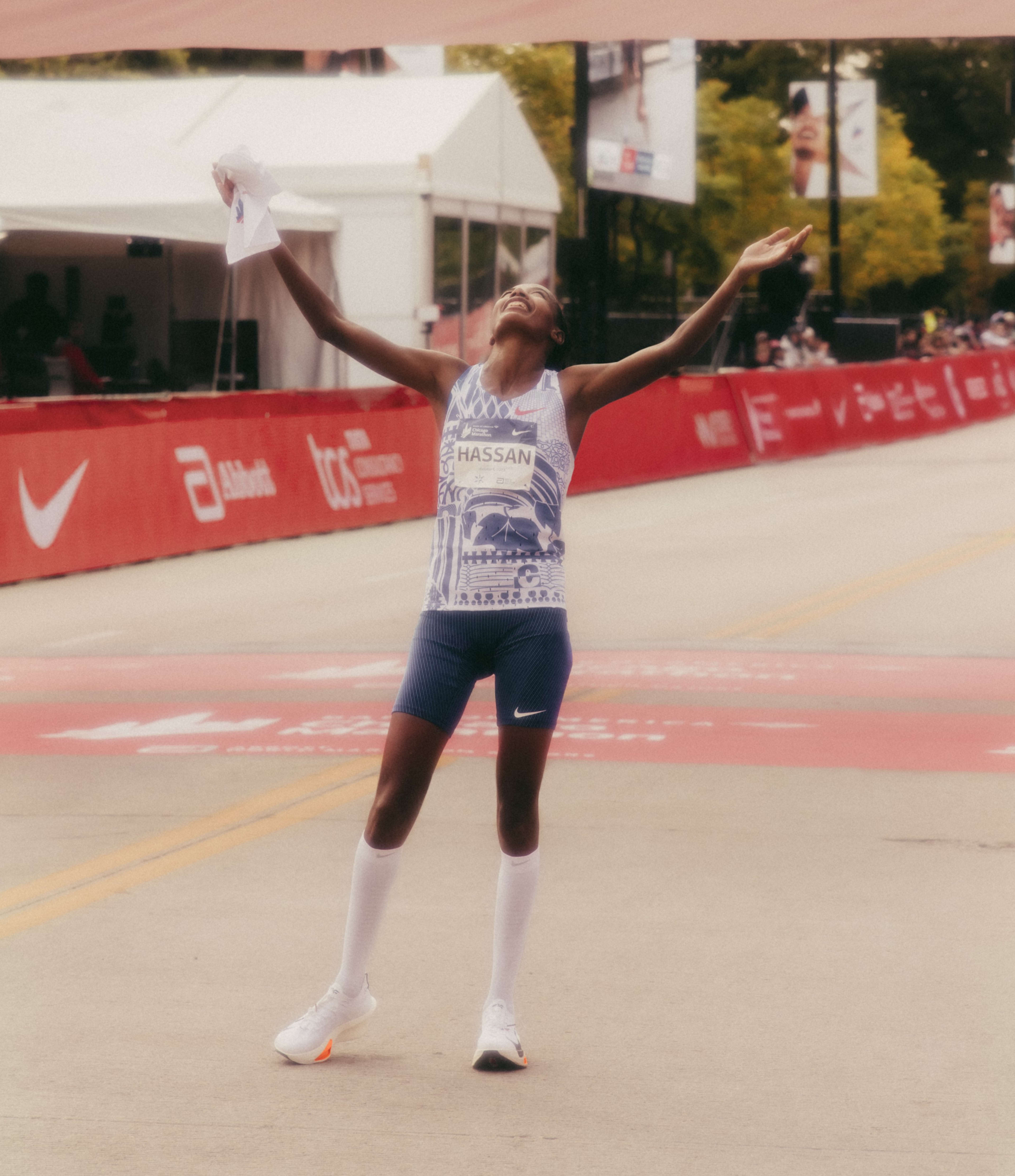 Full body portrait of Sifan Hassan holding her arms up in victory after winning a race. Sifan is in Nike running shoes, high white socks, blue running shorts, and a blue and white patterned singlet.