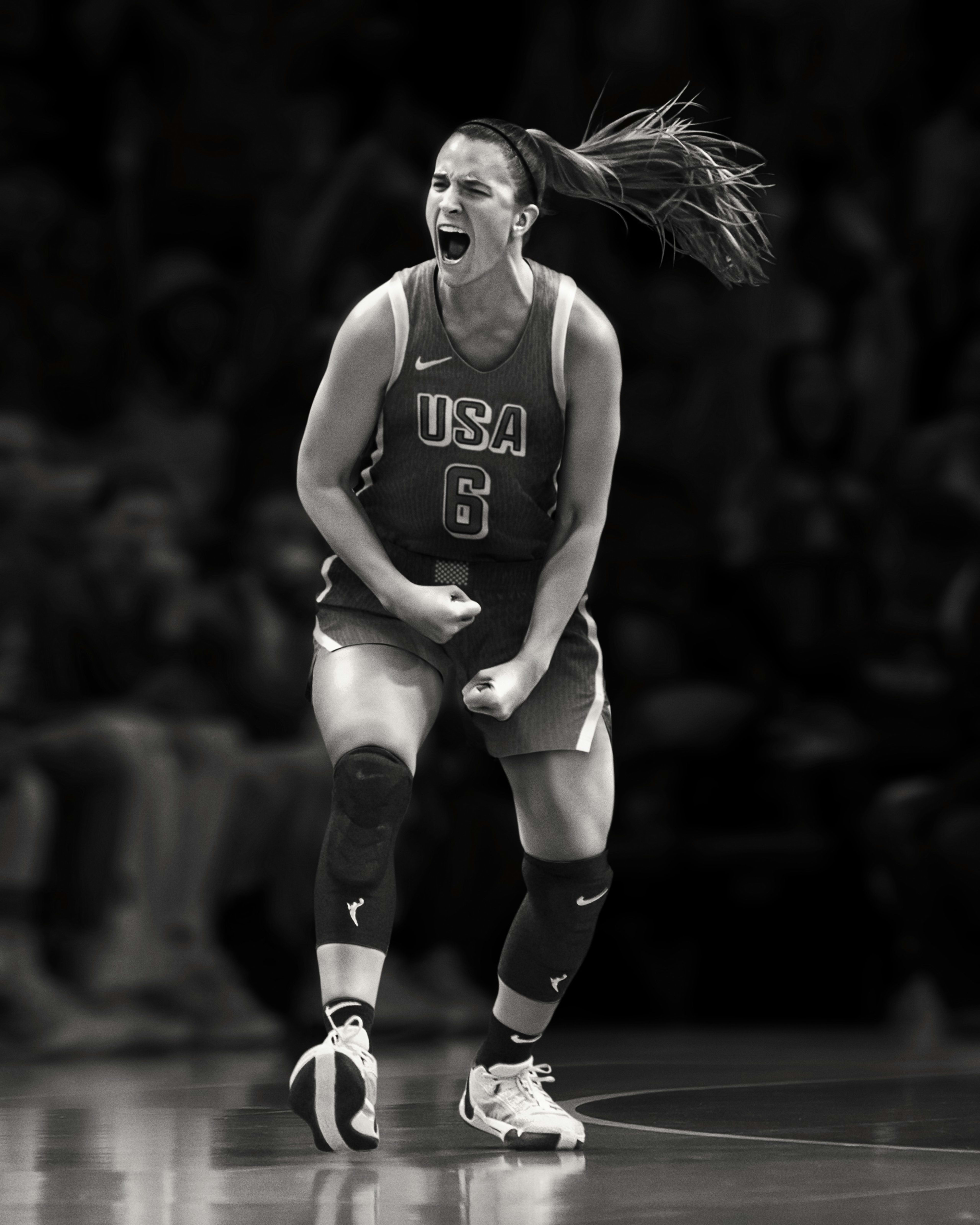 Black and white full body image of Sabrina Ionescu celebrating on the basketball court. She wears her Team USA uniform and her hair is flying out behind her.