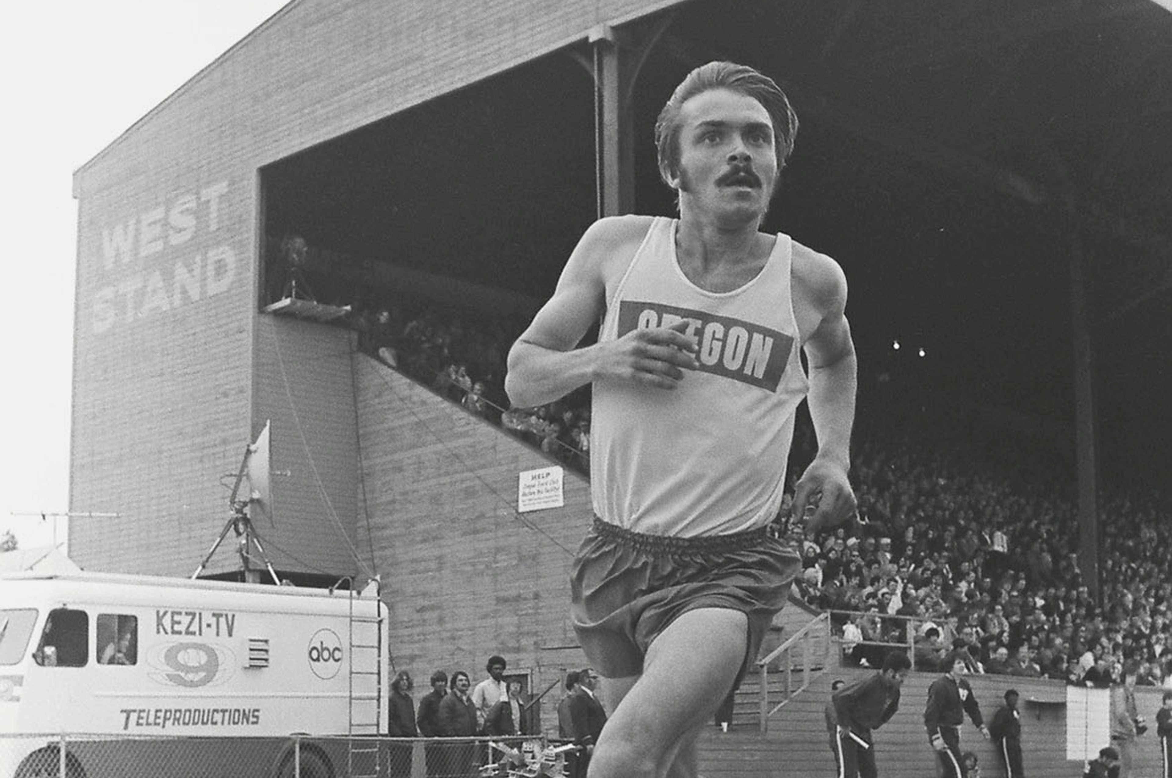 Black and white photo of Steve Prefontaine running on the track at Hayward Field at University of Oregon, Eugene, OR.  Pre wears an Oregon running singlet with dark running shorts and looks down the track as he pumps his arms during a race.