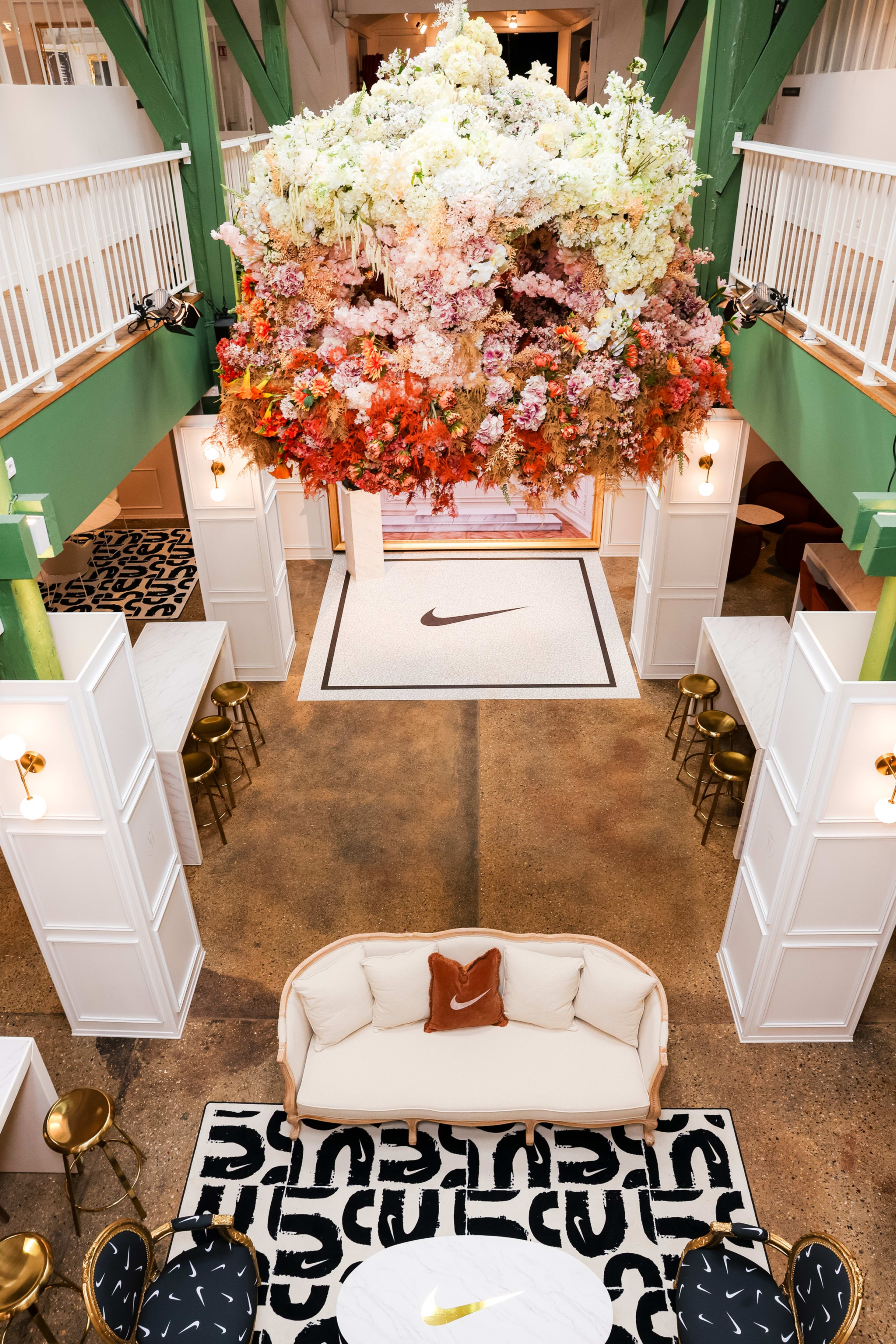 Overhead view into the entryway of the Nike Athlete House in Paris, France.  A dramatic flower display hangs above a lounge space with green walls. 2 chairs are upholstered with black fabric with white Swooshes. A white sofa holds a rust colored pillow with a white Swoosh. There is white cabinetry and high tables with barstools flanking the room.