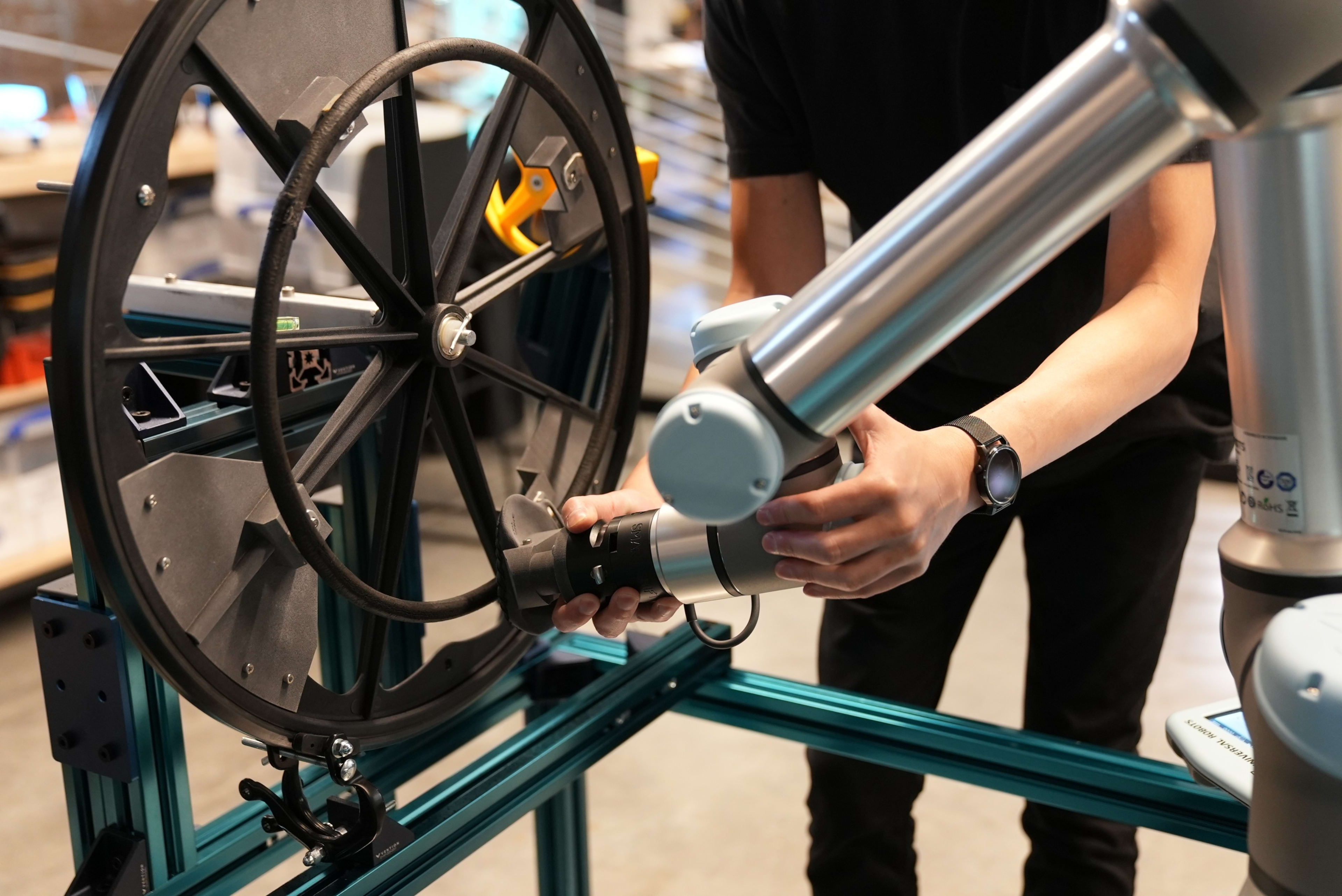 A member of the research team adjusts the robot mechanical arm during testing in the Nike Sports Research Lab, in Beaverton, OR.  We see a close up of  two hands adjusting the silver arm against the black wheelchair wheel.