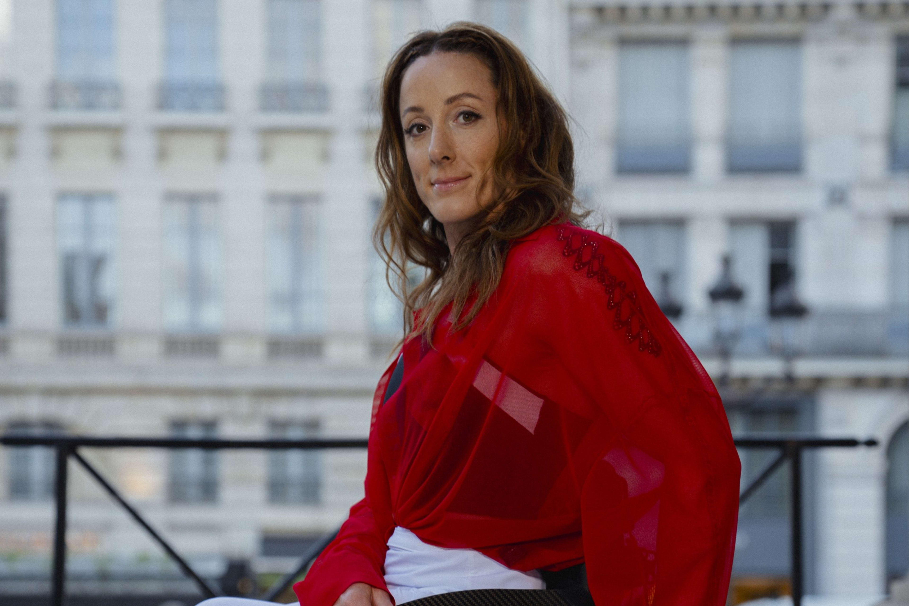 Fully body portrait of Tatyana McFadden sitting in her wheelchair on a marble entryway with European buildings in the background. Tatyana wears a red blouse, white track pants, and Nike Pegasus EasyOn shoes. She smiles towards the camera.