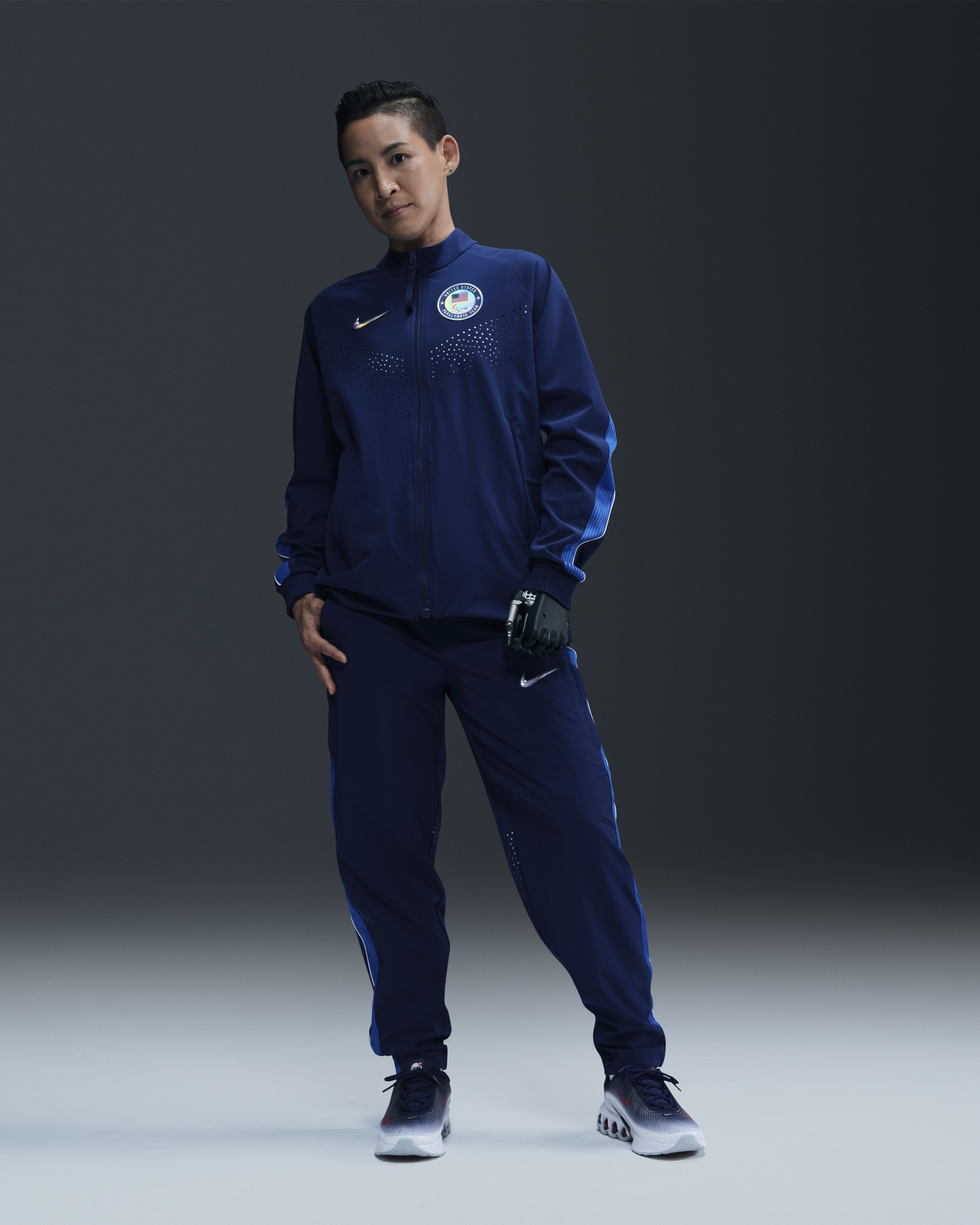 Full body view of a female para-athlete wearing the Women's Medal Ceremony Jacket, Pant, and Nike Air Max Dn footwear, all in the exclusive bright blue Team USA colorway. The athlete stands on a white floor against a grey background.