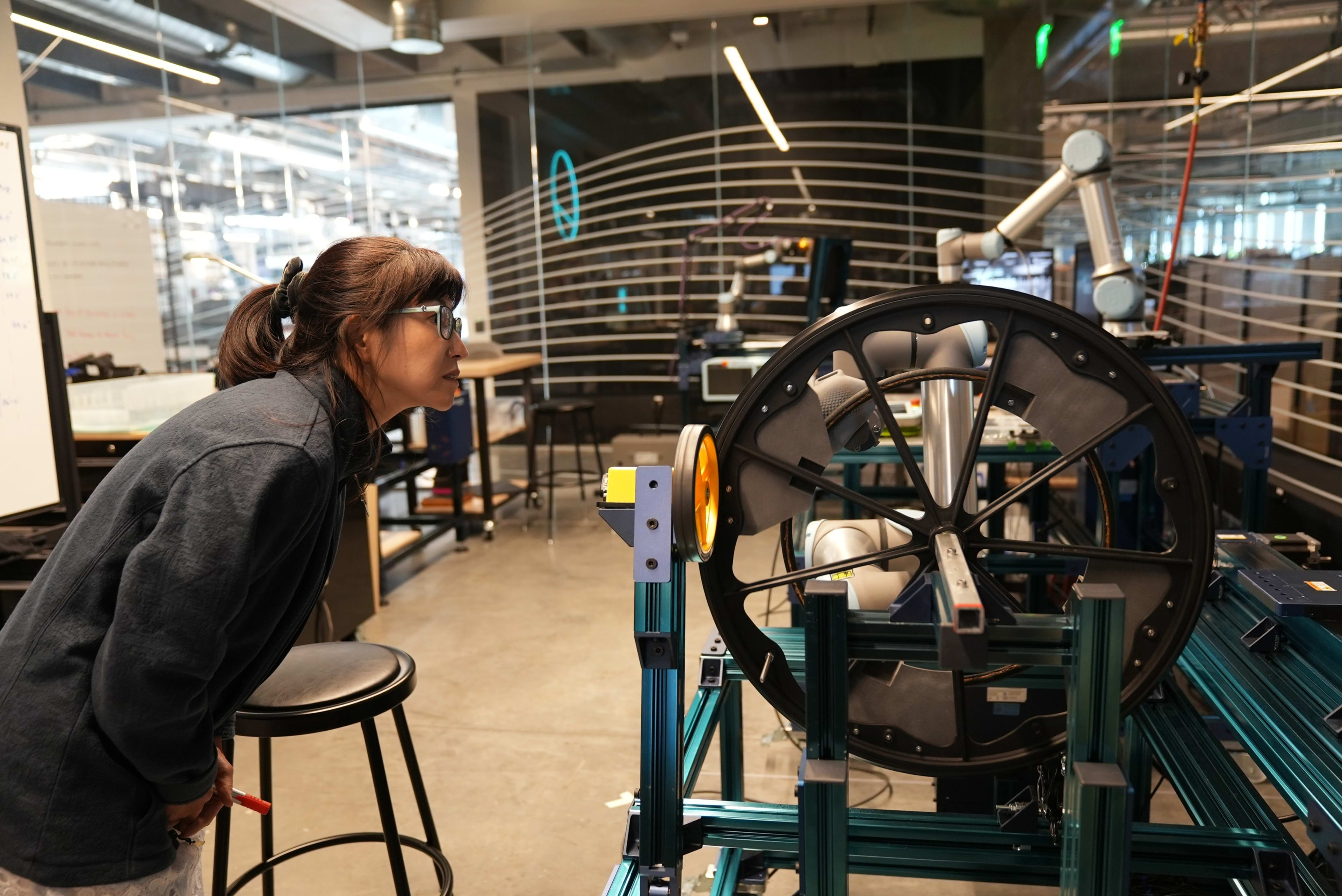 Eunice Lee leans in to review the robot arm and wheel testing underway in the Nike Sports Research Lab, in Beaverton, CA. Eunice looks right at the wheel, which sits in a lab space with tables, chairs, and easels in the background.