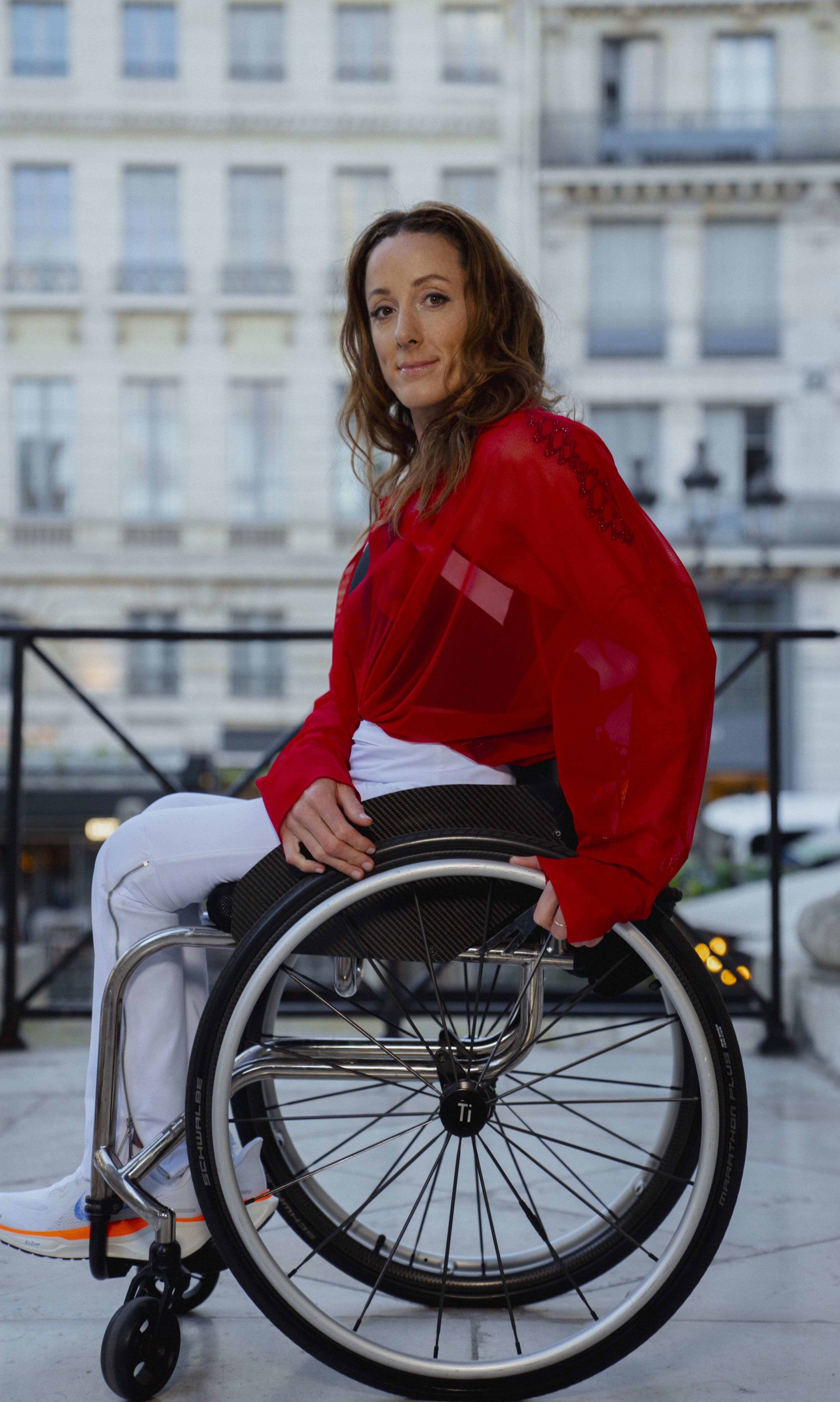 Fully body portrait of Tatyana McFadden sitting in her wheelchair on a marble entryway with European buildings in the background. Tatyana wears a red blouse, white track pants, and Nike Pegasus EasyOn shoes. She smiles towards the camera.