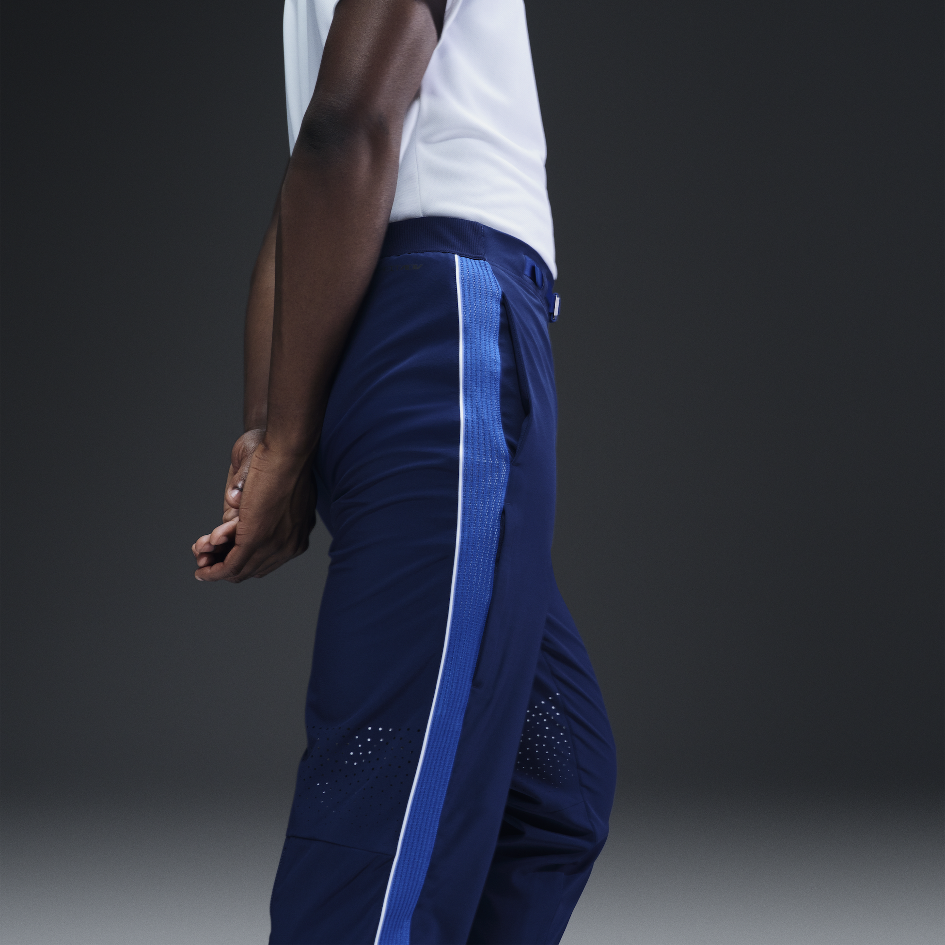 Side view of a male athlete wearing the Men's Medal Ceremony Pant in the exclusive bright blue Team USA colorway. He has a white Nike tee tucked into the pant, while he stands with hands linked behind his back against a dark background.