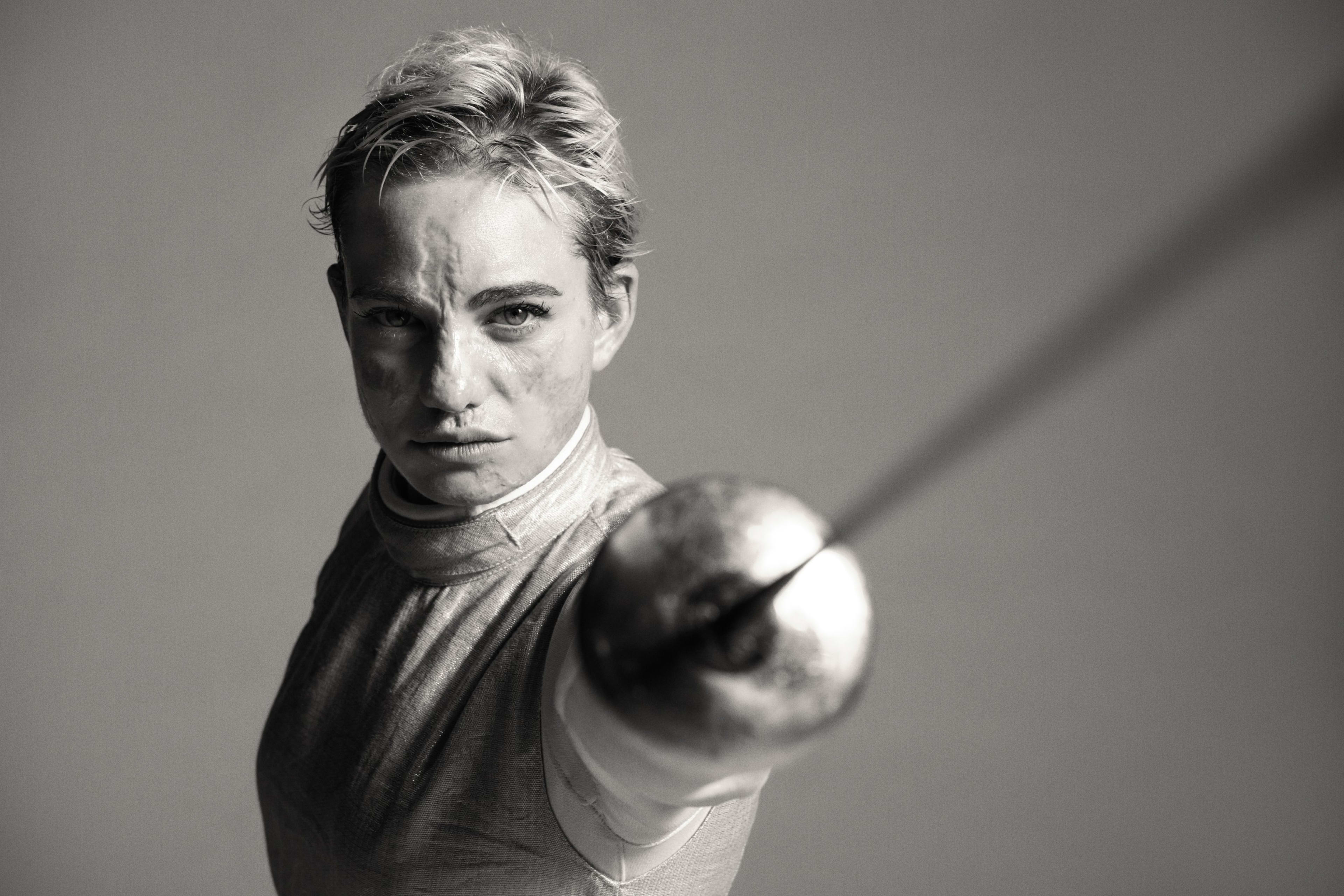 Black and white front angled view of Bebe Vio thrusting a fencing foil forward in a stanced position.