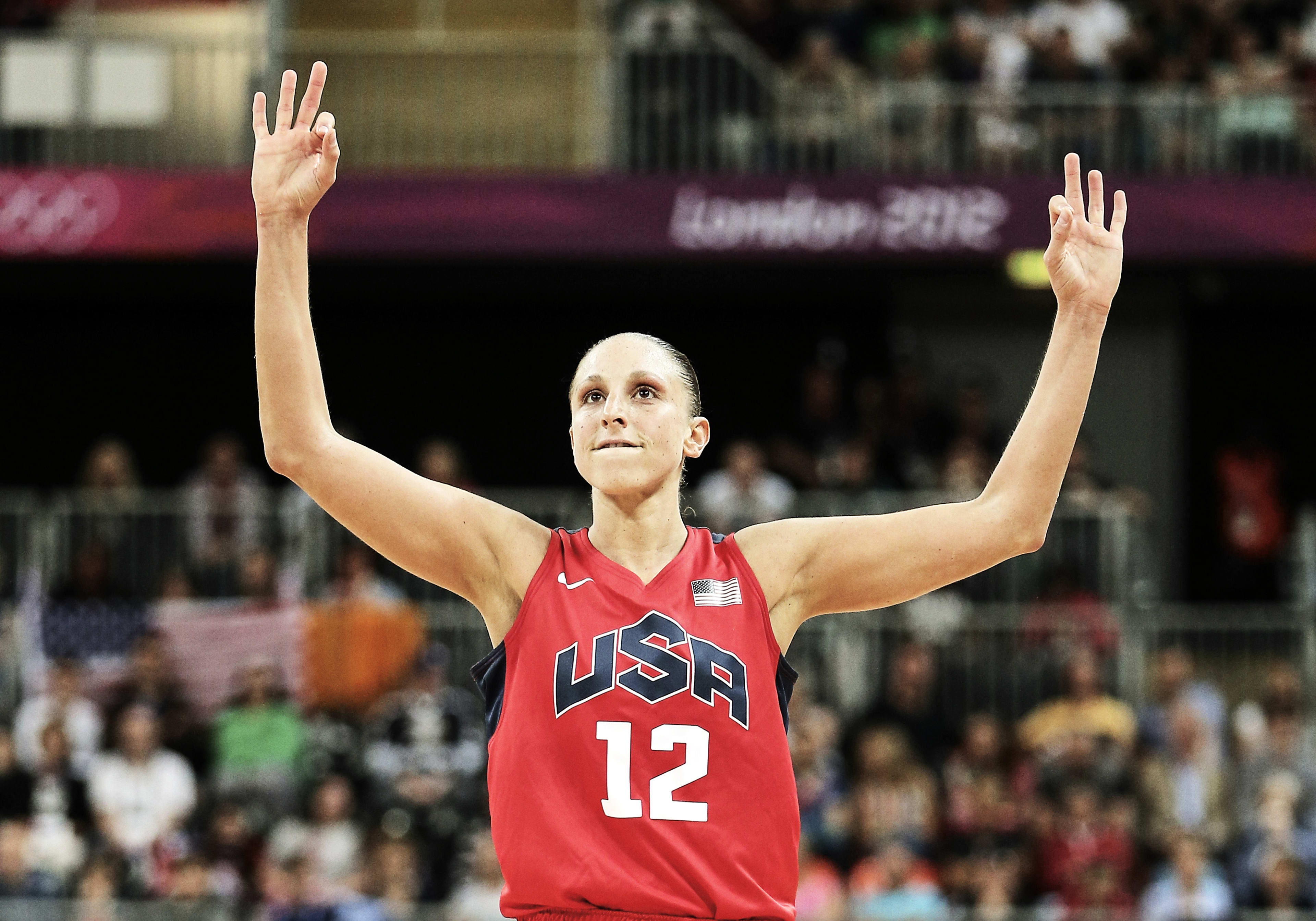 Diana Taurasi holds her hands high, with six fingers raised, signaling her 6 gold medals. Diana wears a read Team USA jersey, and a crowd looks on behind her.