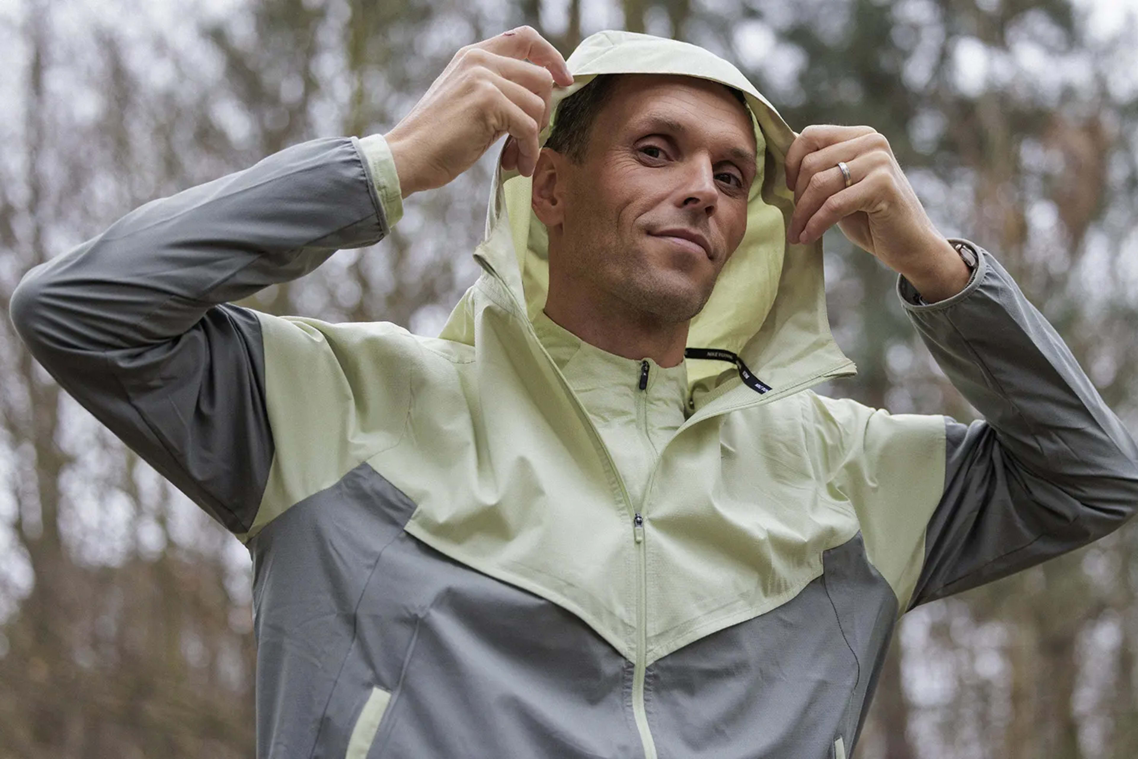 Alexis Hanquinquant pulls the hood of his Nike Windrunner Repel jacket up over his head. He smiles towards the camera, and we can see trees in the background.