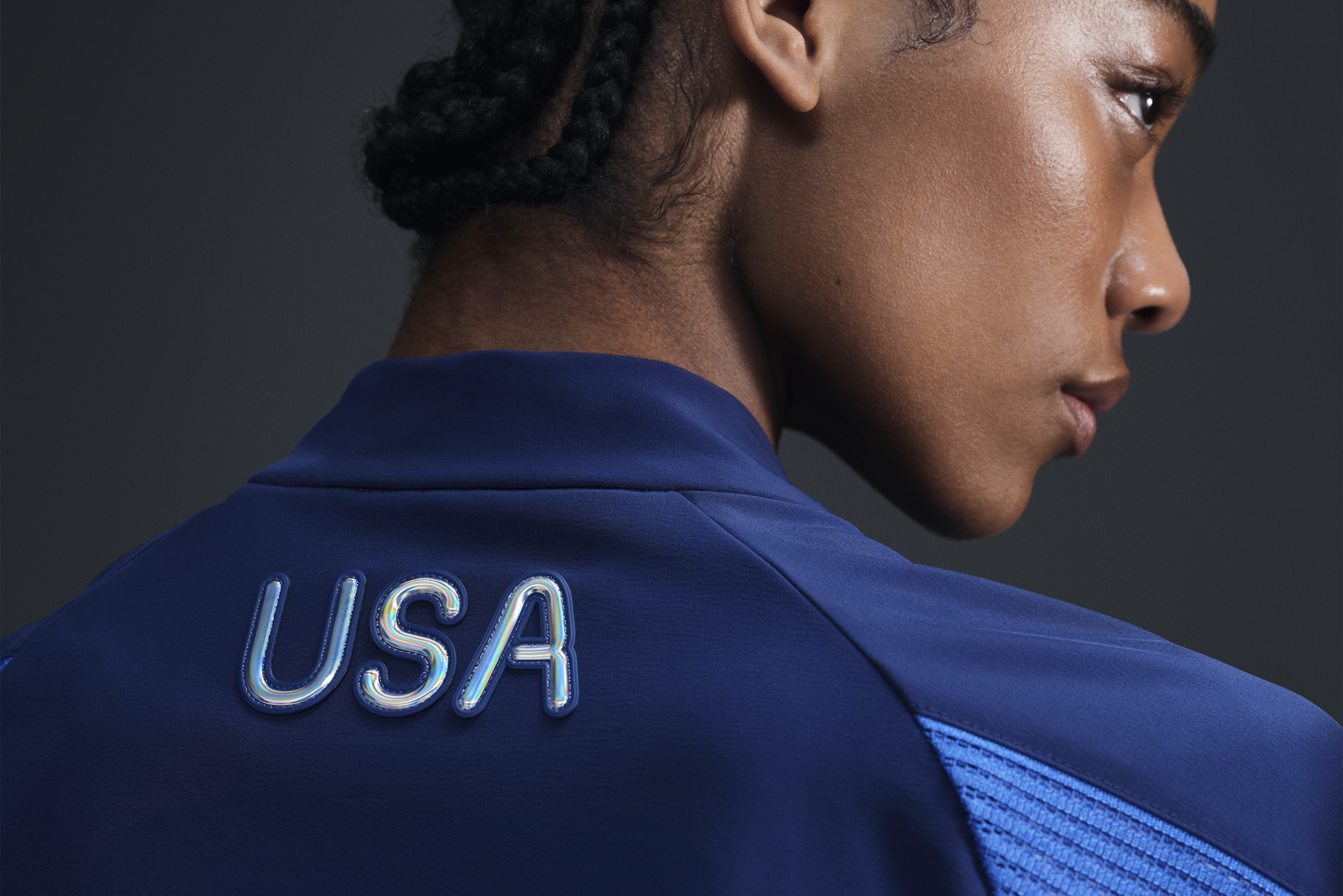 Back view of athlete wearing Women's Medal Ceremony Jacket in the exclusive bright blue Team USA colorway.  USA is emblazoned on the back of the jacket in iridescent silver letters. She stands against a dark background.