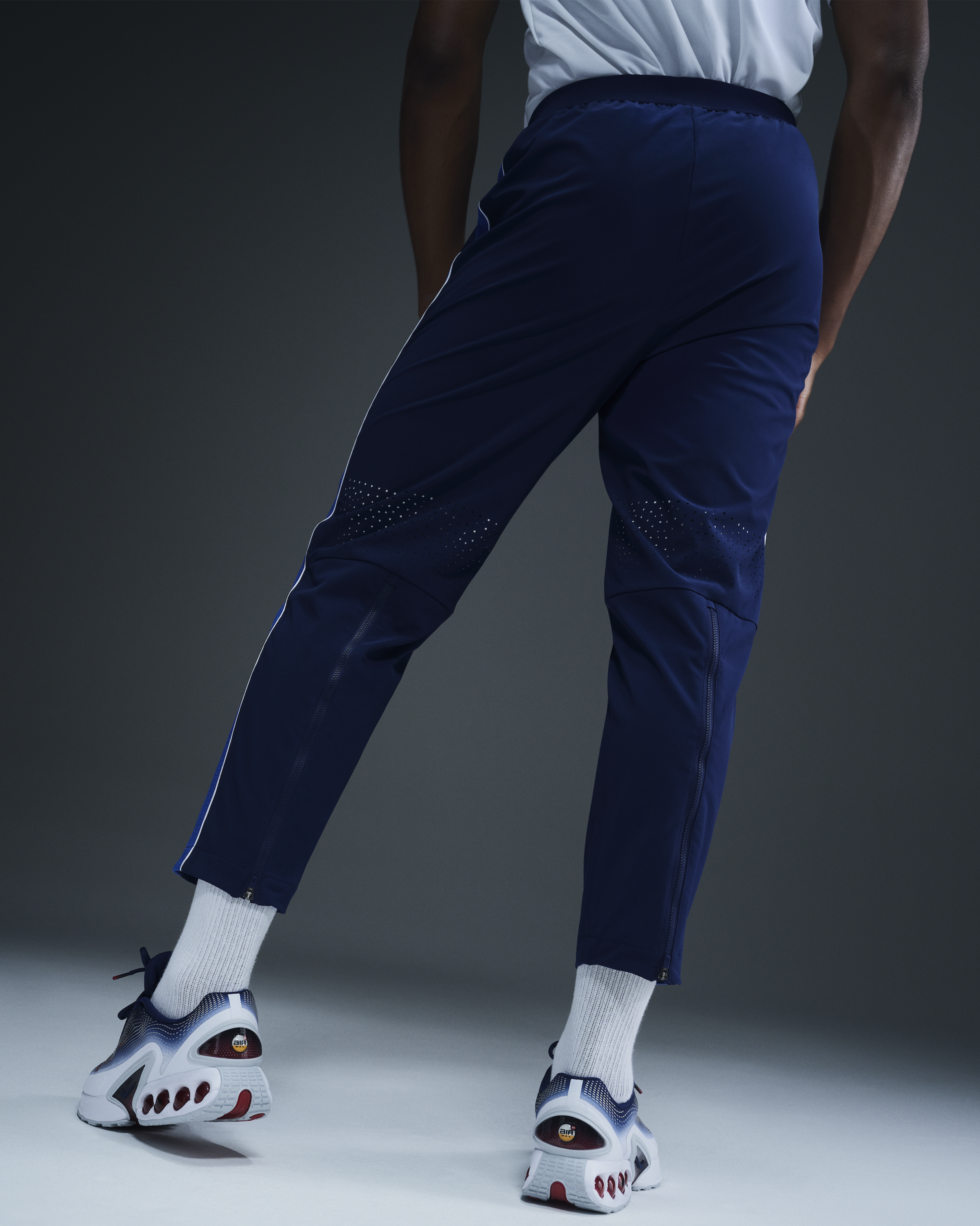 Back, waist-down view of a male athlete wearing the Men's Medal Ceremony Pant in the exclusive bright blue Team USA colorway. He wears white socks with the Team USA Nike Air Max Dn footwear. The athletes stands on a white floor against a dark grey background.