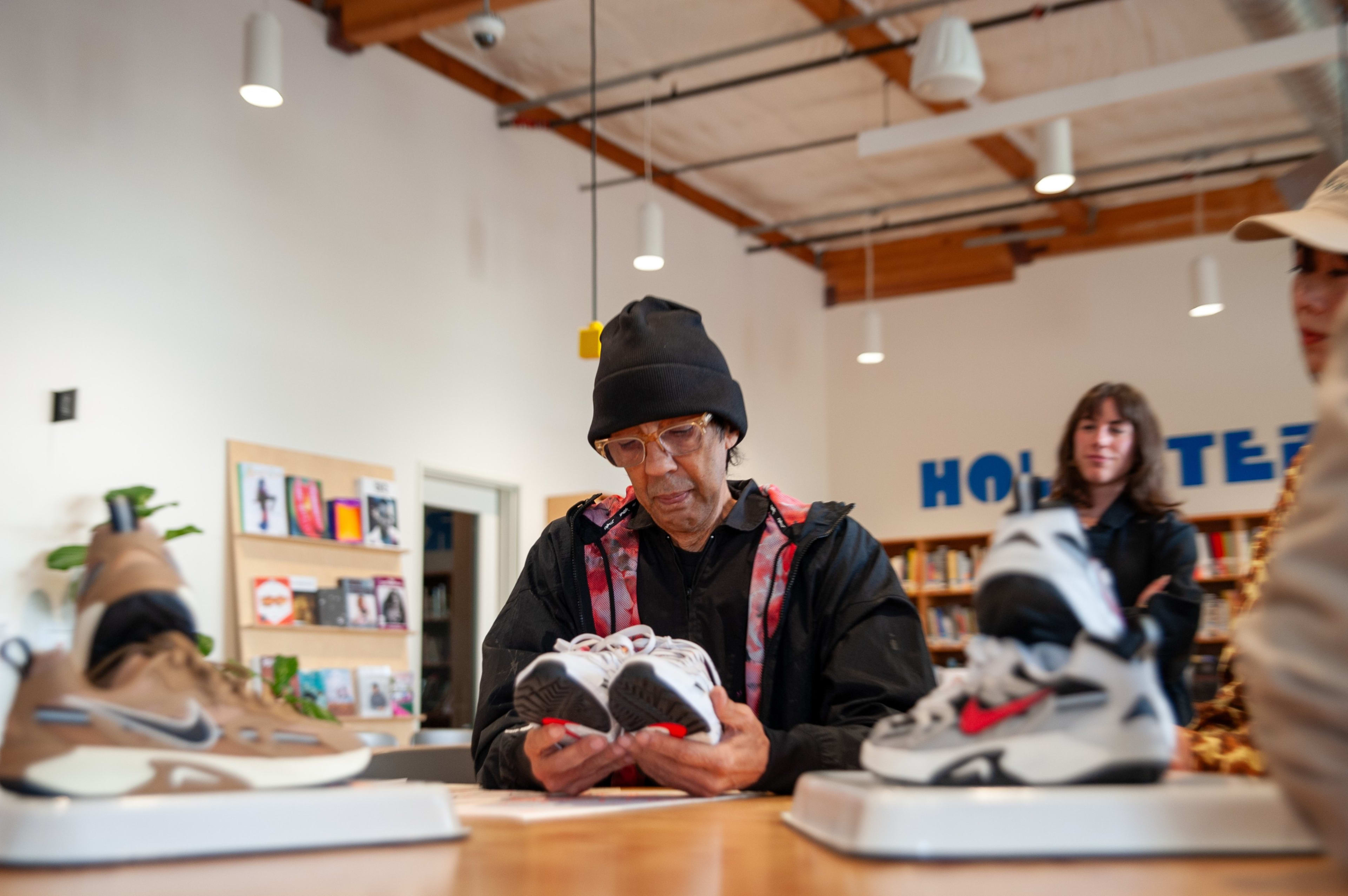 Futura sits at a table in Nike's Blue Ribbon Studio in Beaverton, Oregon. Futura wears a black beanie hat, clear framed glasses, and the Nike X Futura hoodie in black. He reviews Nike Jam sneakers in white and tan colorways.