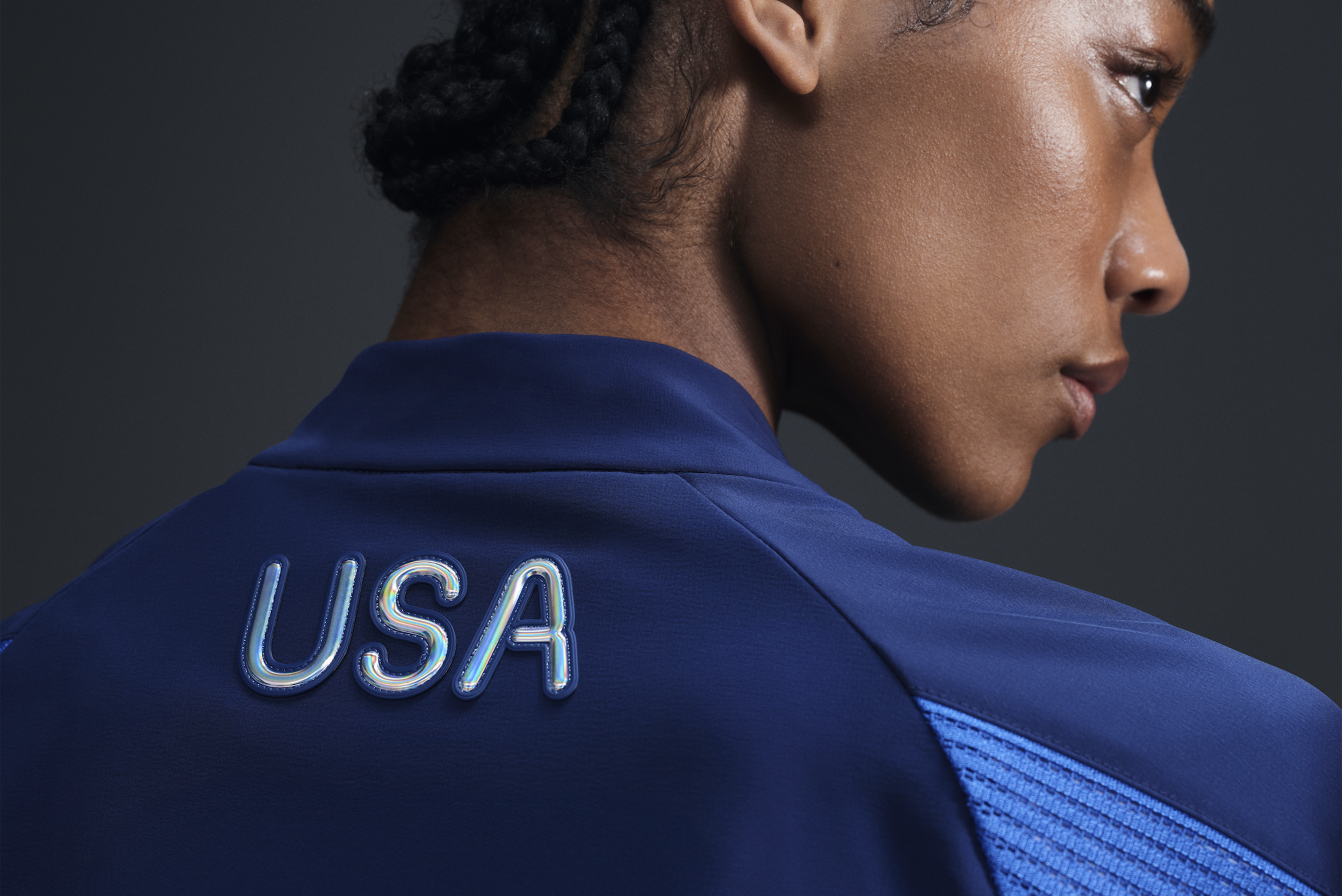 Back view of athlete wearing Women's Medal Ceremony Jacket in the exclusive bright blue Team USA colorway.  USA is emblazoned on the back of the jacket in iridescent silver letters. She stands against a dark background.