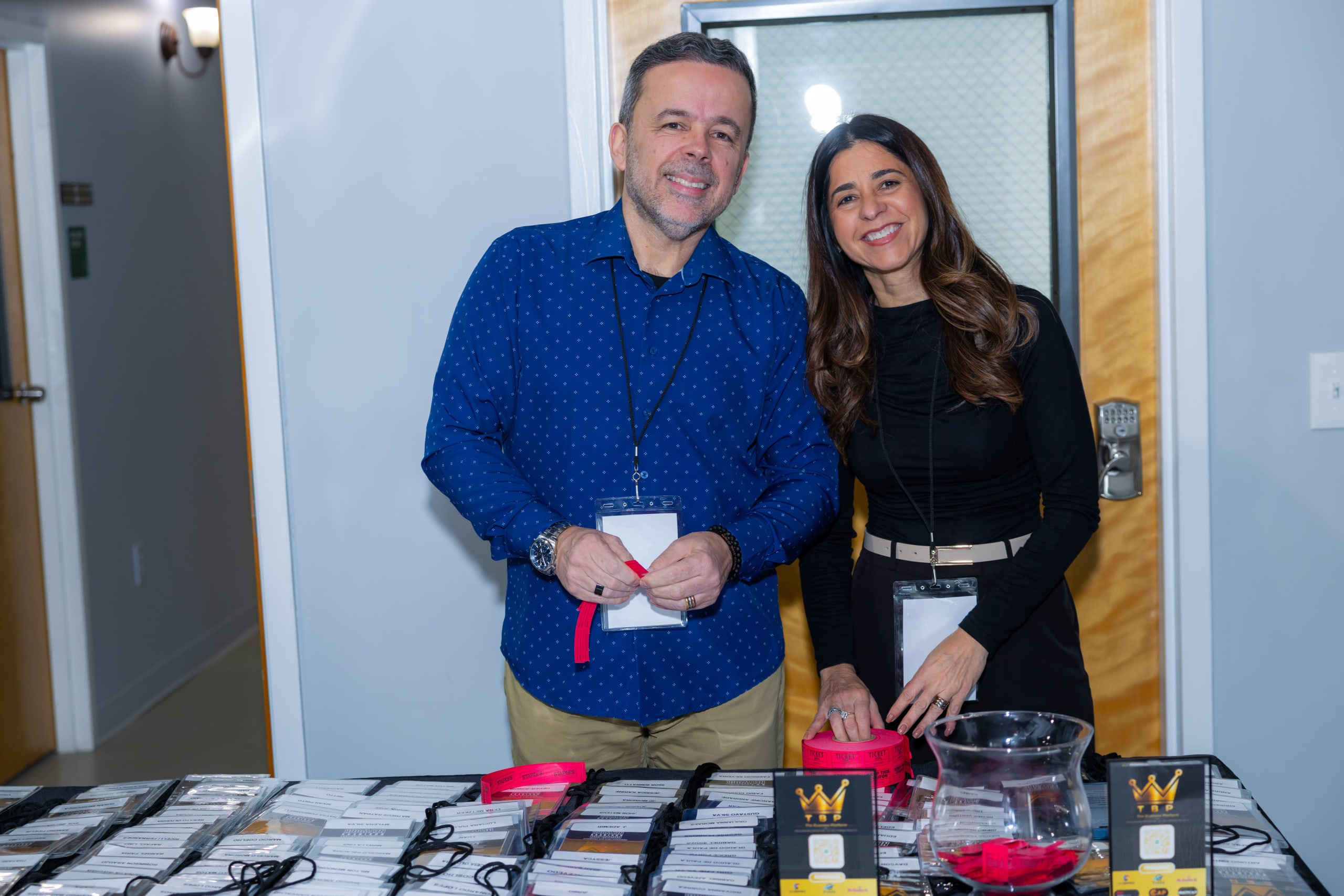 Um homem e uma mulher sorriem na mesa de credenciamento, ao lado dos crachás do BBN