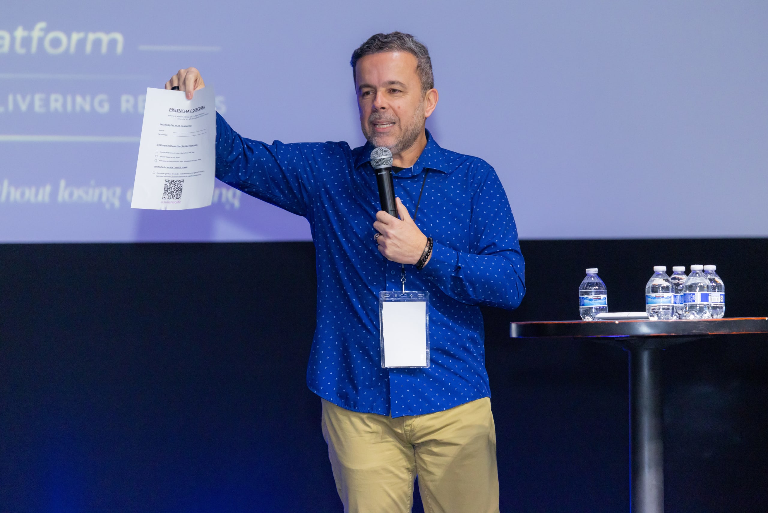 Palestrante no palco do Encontro BBN apresenta ao microfone, mostrando ao salão uma folha com QR code