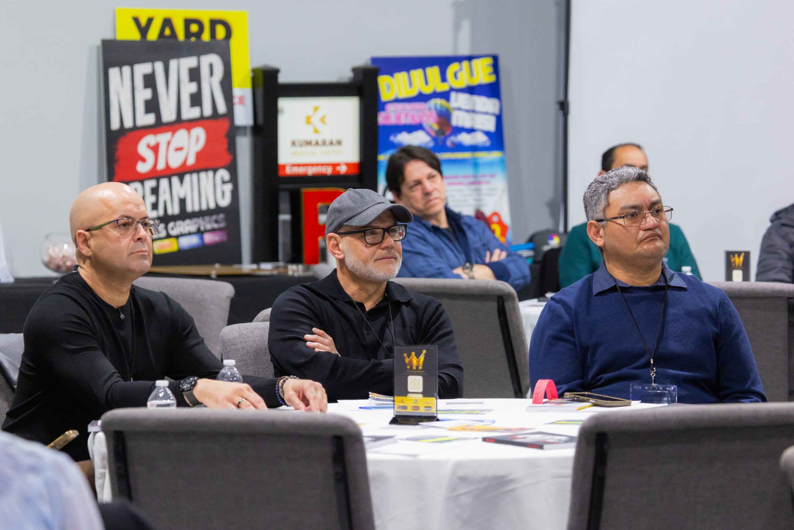 Participantes acompanham uma palestra sentados à mesa, com banners de patrocinadores ao fundo