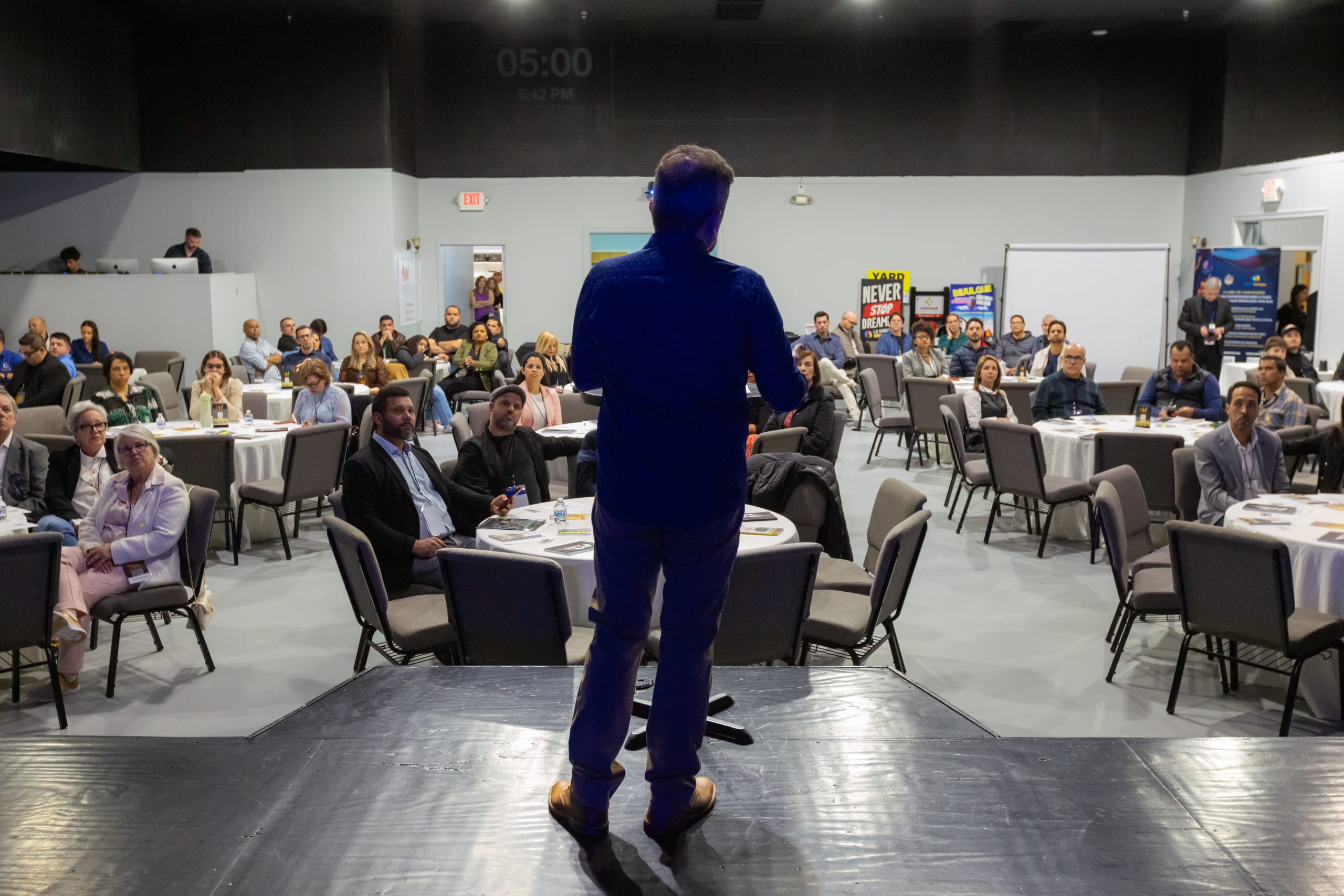 Palestrante no palco, visto de costas, fala para o salão cheio de participantes em mesas redondas
