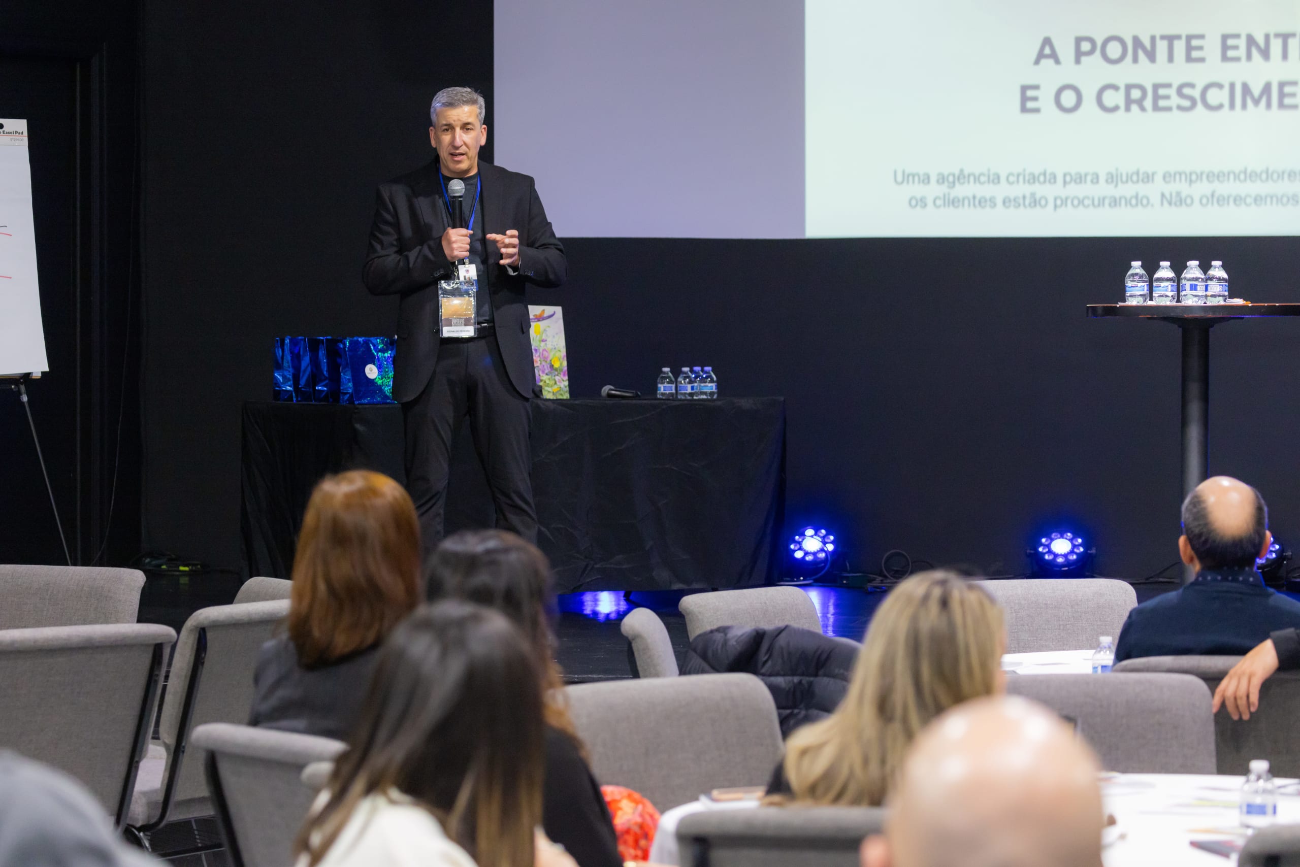 Palestrante fala ao microfone no palco enquanto o público acompanha em primeiro plano