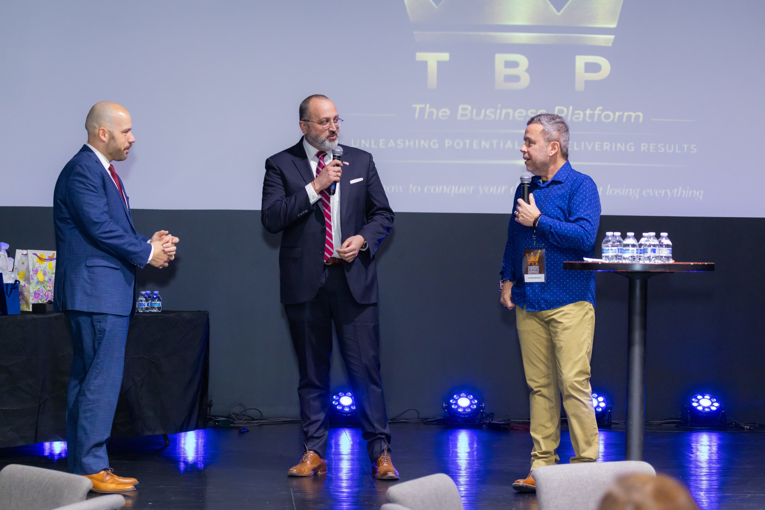 Três homens conversam no palco, dois deles com microfone, diante do telão do evento