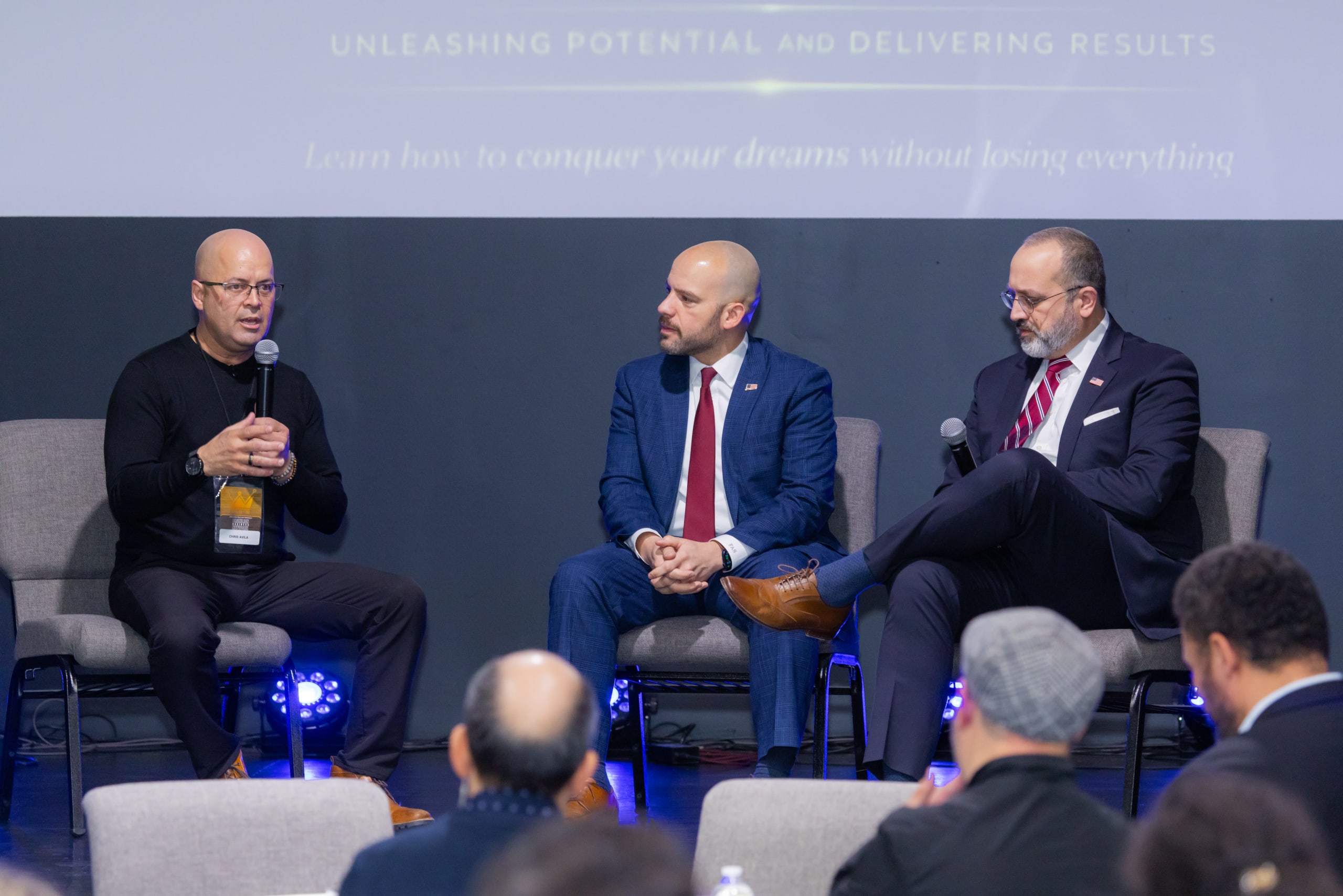 Painel com três convidados sentados no palco, observados pelo público em primeiro plano