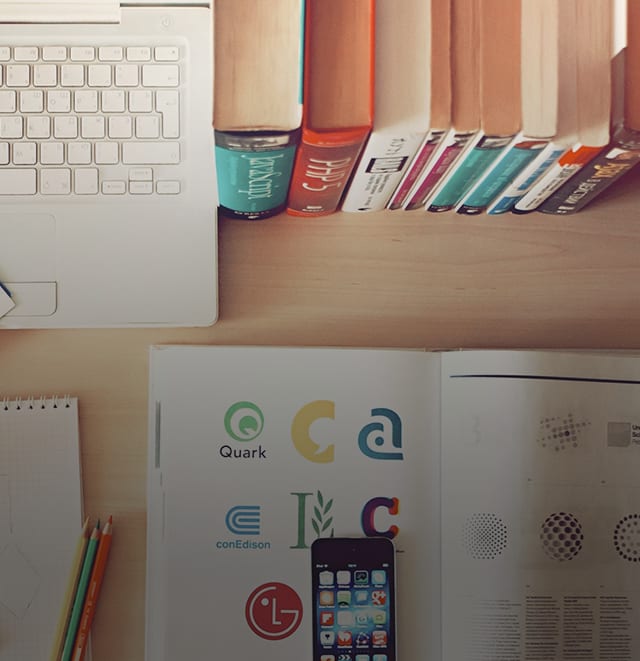 A logo design book lying open on a computer desk by a computer and other books.