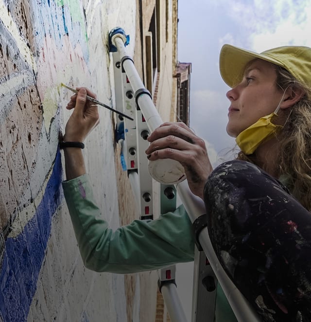 girl on a ladder painting a mural on a brick building with a paint brush