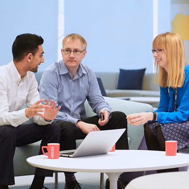 3 Nestle Tech employees talking around a coffee table, drinking coffee.