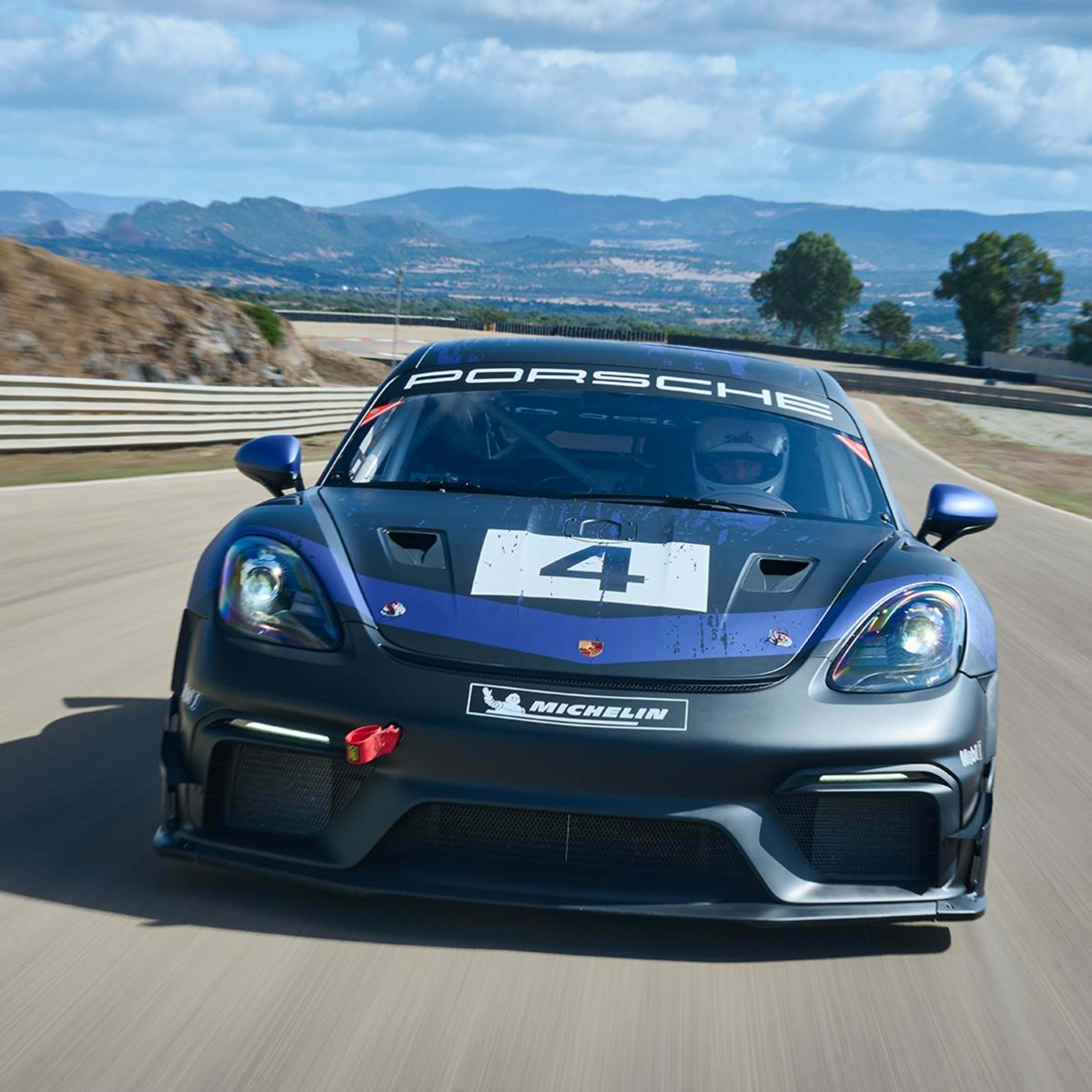 the blue 718 Cayman GT4 RS Clubsport racing on the track with mountains on each side and seen from the front