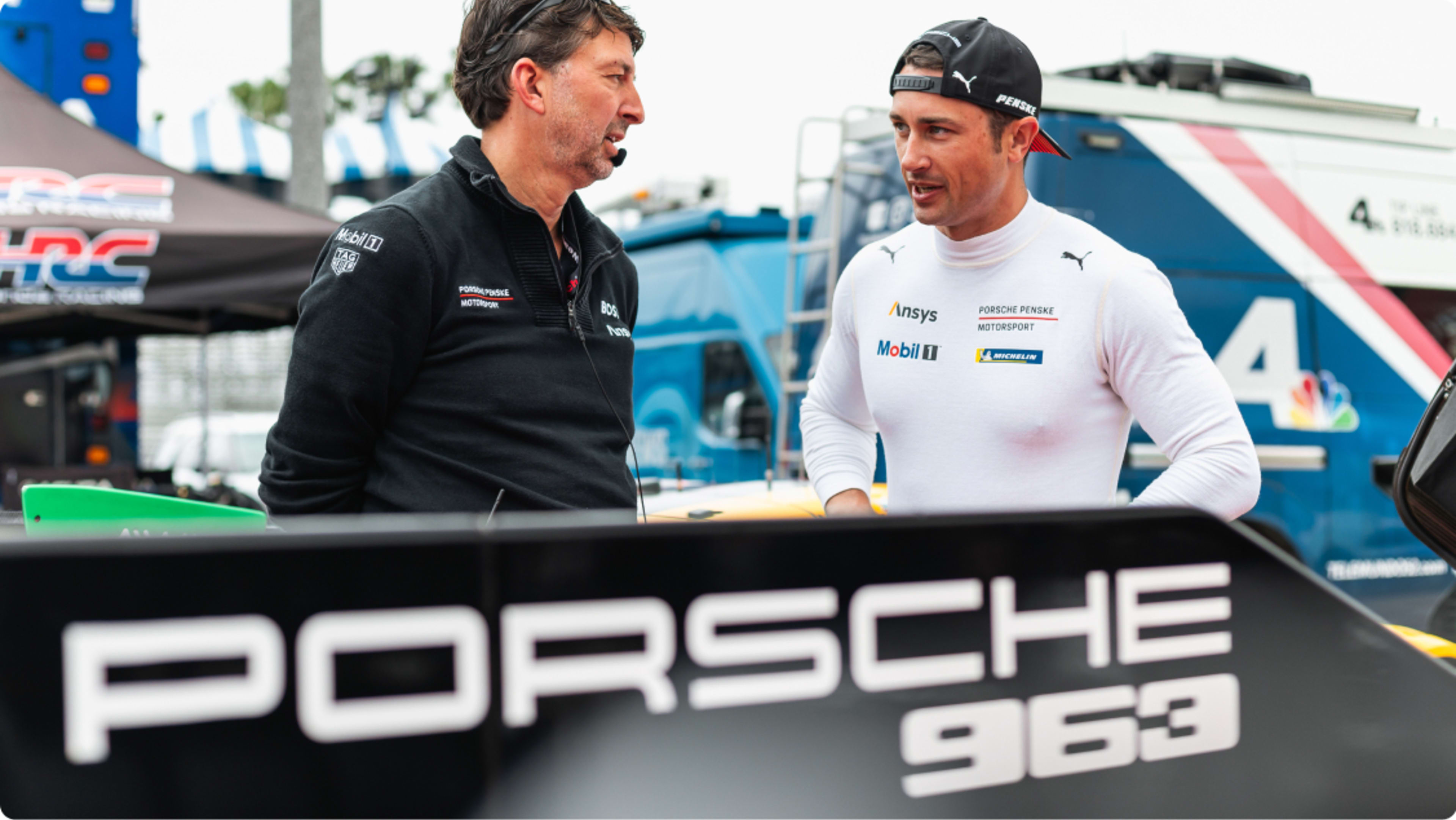 Dane Cameron talking to his colleague behind a Porsche 963 banner 