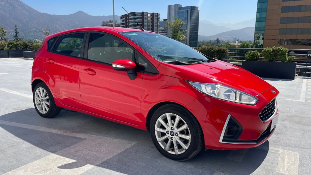 Ford Fiesta SE 2019 rojo manual - Vista 3/4 de hatchback color rojo en exterior con fondo de edificios y montañas