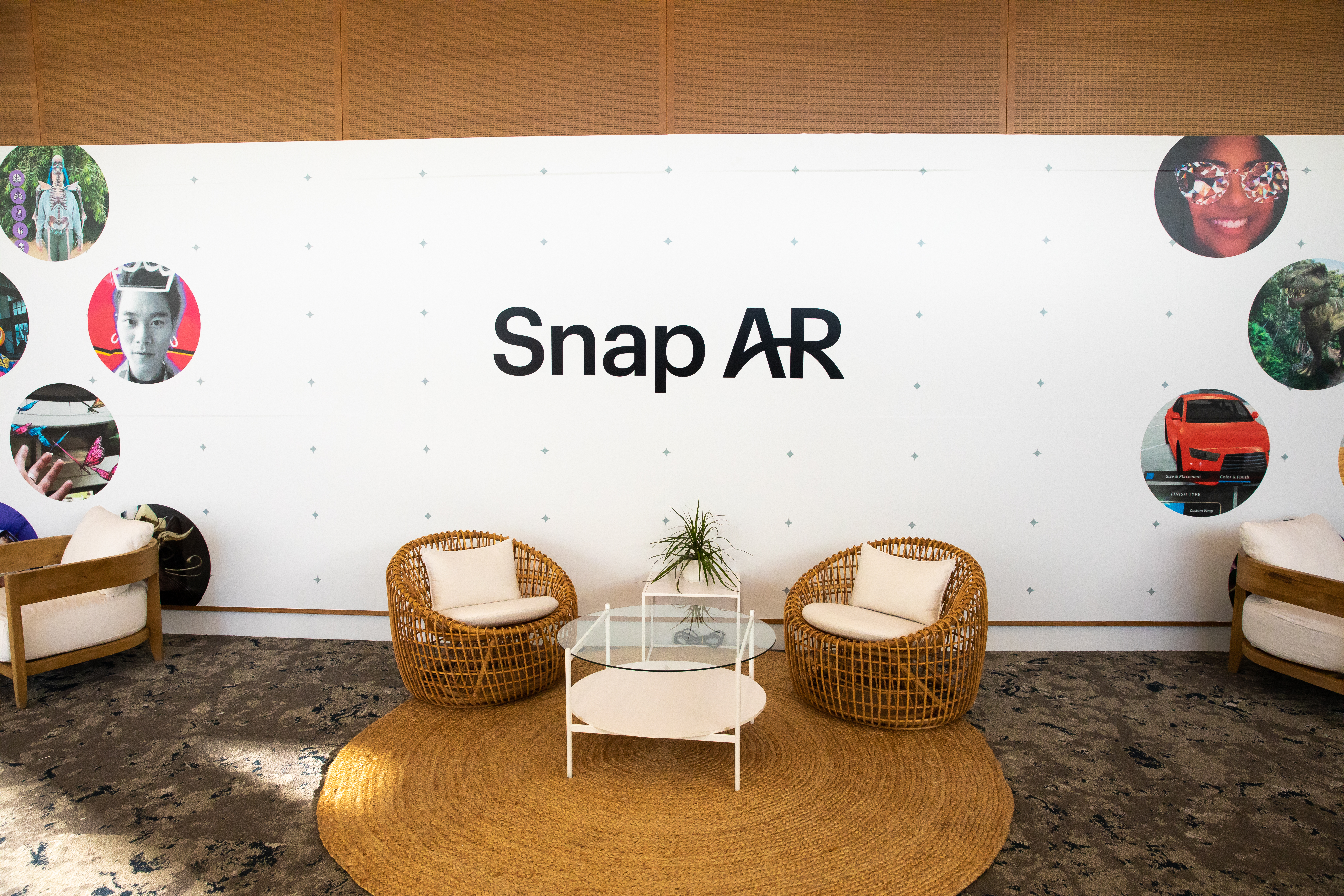 A branded lounge by Snap AR from AWE 2022. two low, modern metal wire chairs in white sit on either side of a large white banner that says "Snap AR." the banner also has many images of 12" circles with photos of peoples faces in them