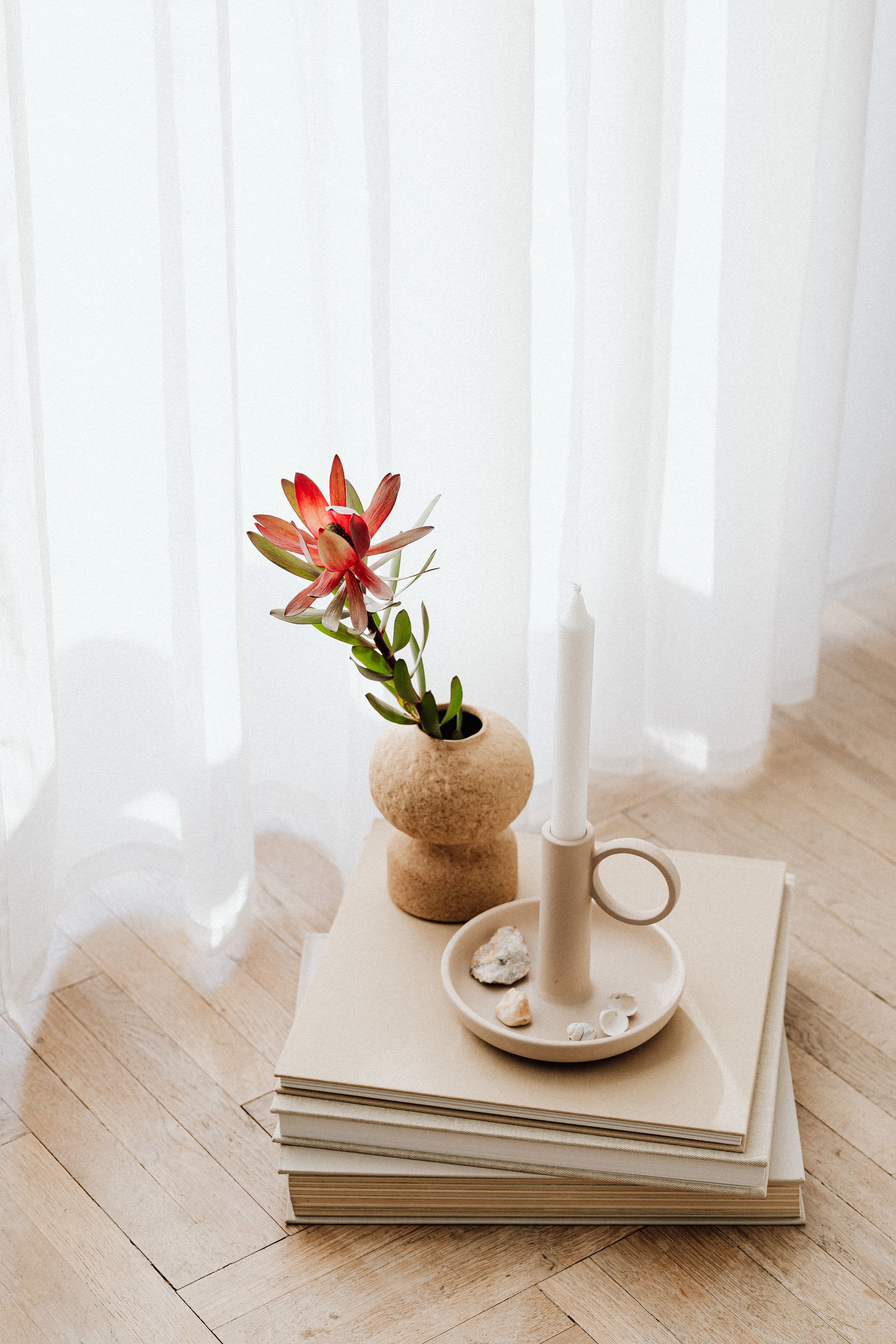 A single flower in a small vase and a candle on a stack of books beside a sheer curtain